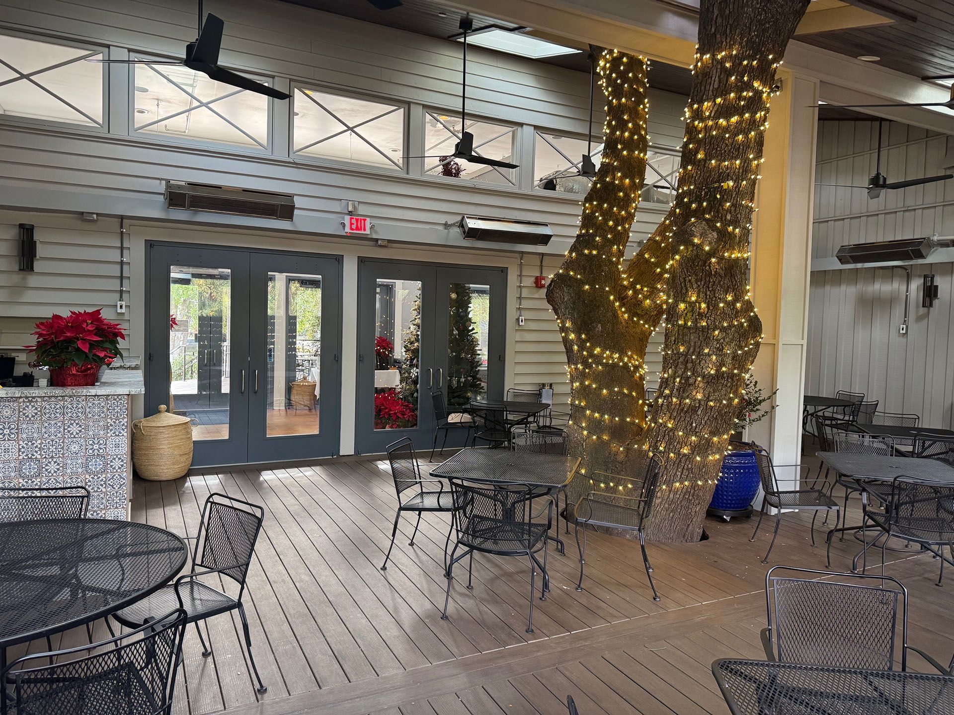 Commercial restaurant remodel and expansion  - Nunzios/After: Outdoor dining area with tables, chairs, and trees wrapped in lights. Double doors lead inside; a bar is to the left.