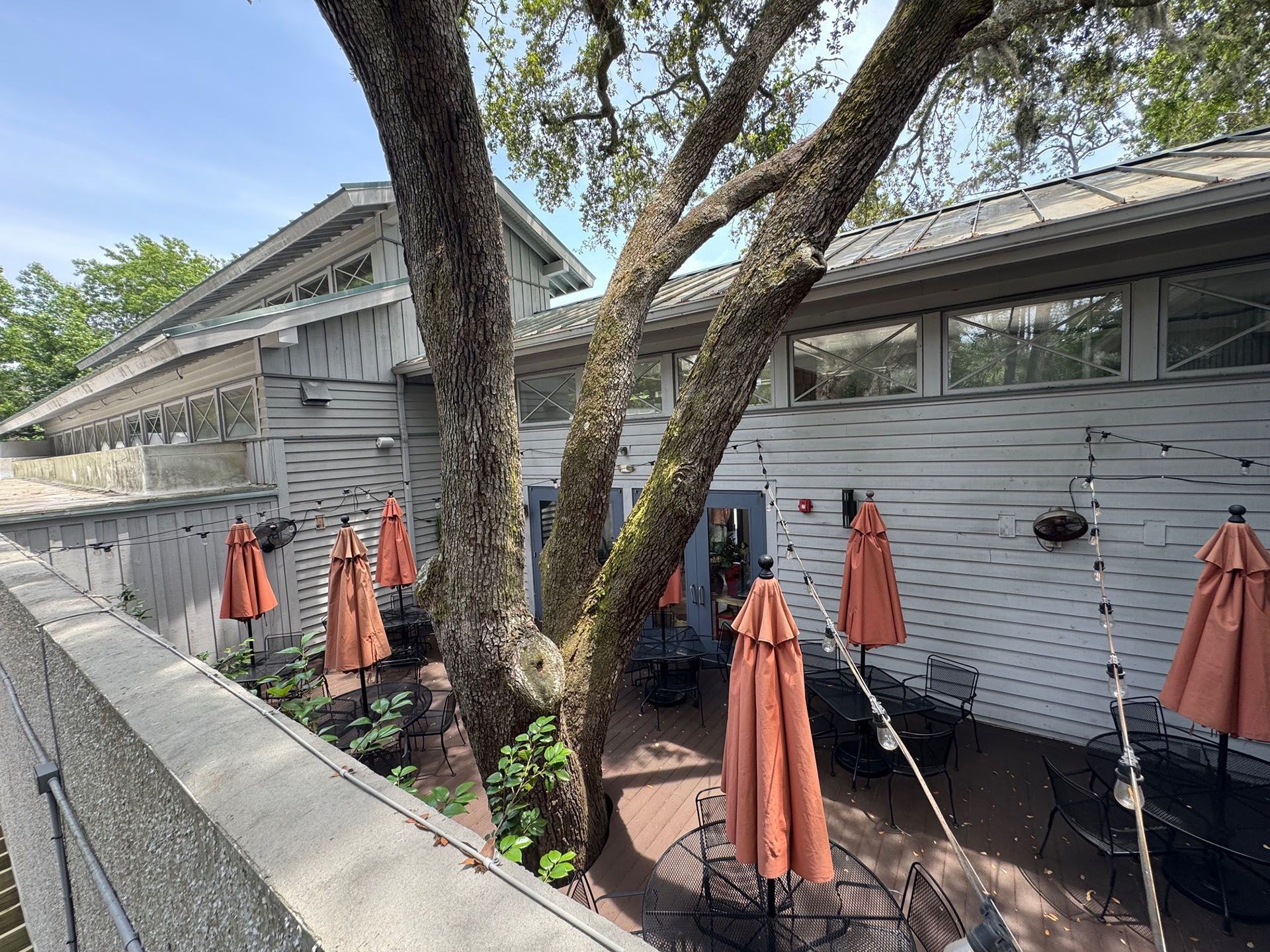 Commercial restaurant remodel and expansion  - Nunzios/Before: Restaurant patio with closed orange umbrellas, tables, and tree branches. White building in background.