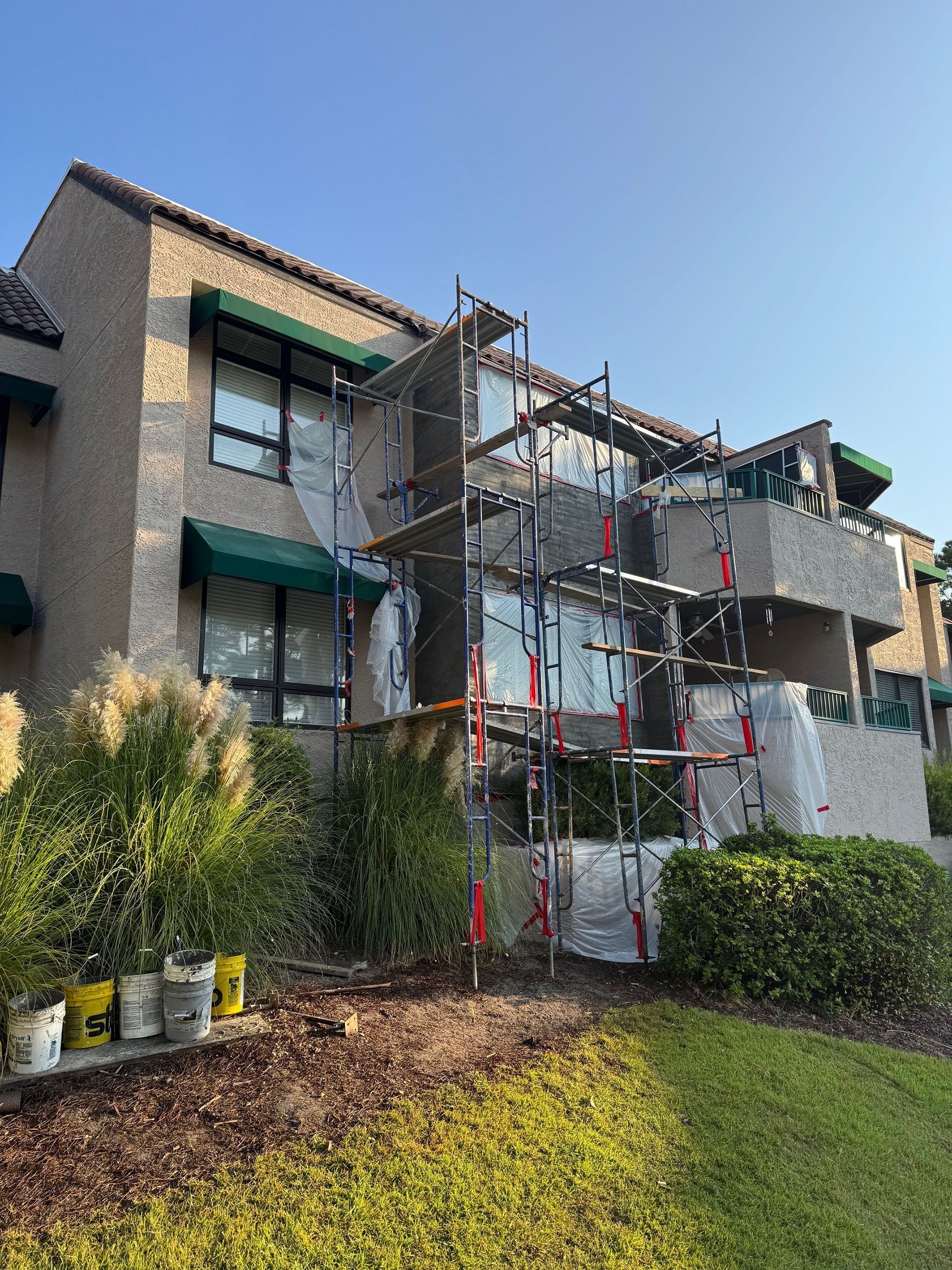 Commercial water damage - During: Scaffolding on building exterior covered in protective material. Green awnings and bushes. Paint buckets sit on the ground.