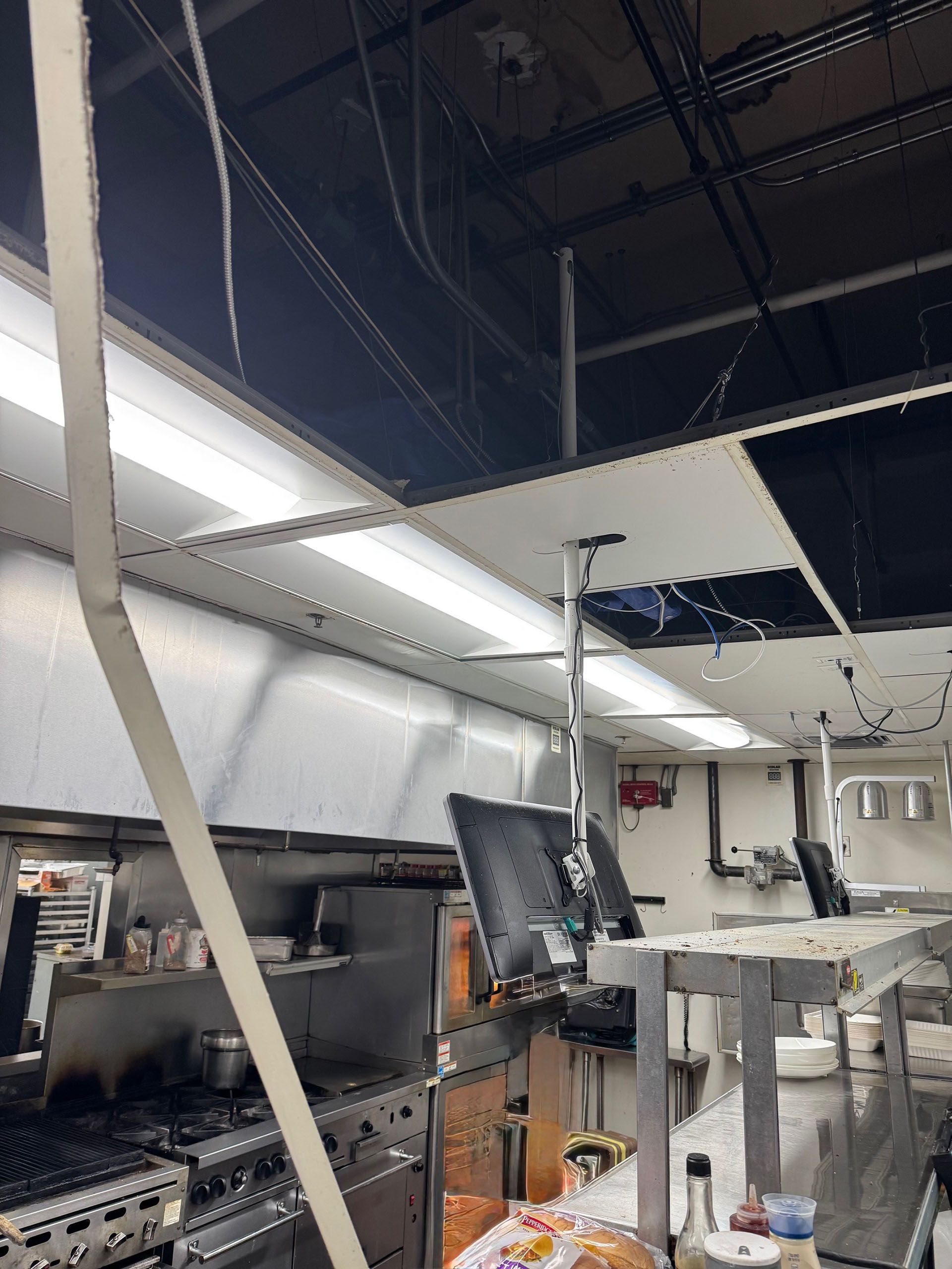 Commercial water damage - Before: Damaged kitchen ceiling, exposed wires, and missing tiles. Stainless steel counters and appliances visible.