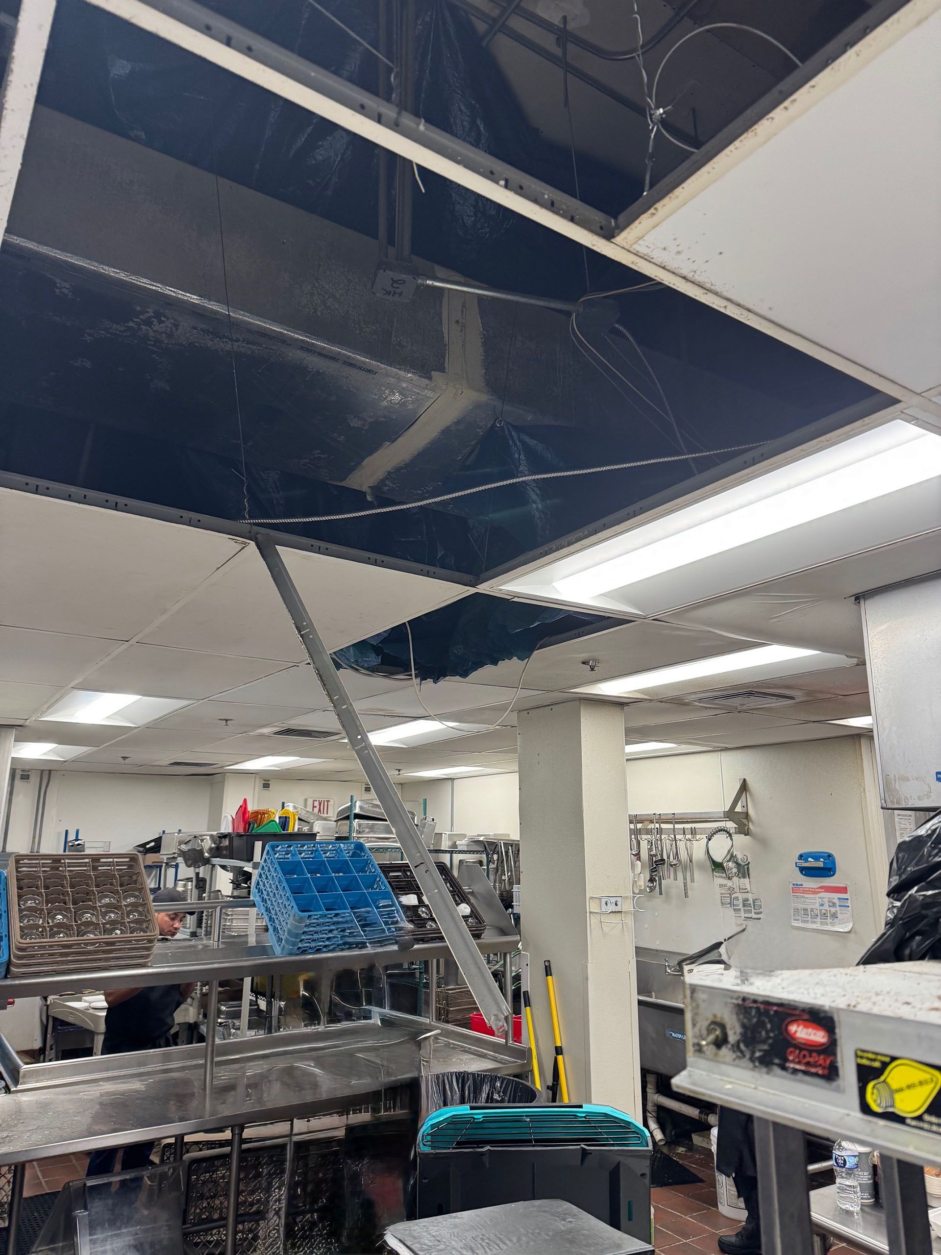 Commercial water damage - Before: Damaged ceiling in a commercial kitchen. Black tile hanging down, supported by a pole. Interior shot.