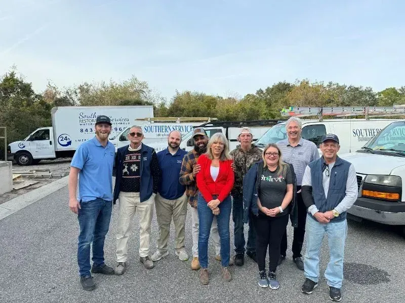 A group of people posing in front of work vans. Trees in the background, blue sky.