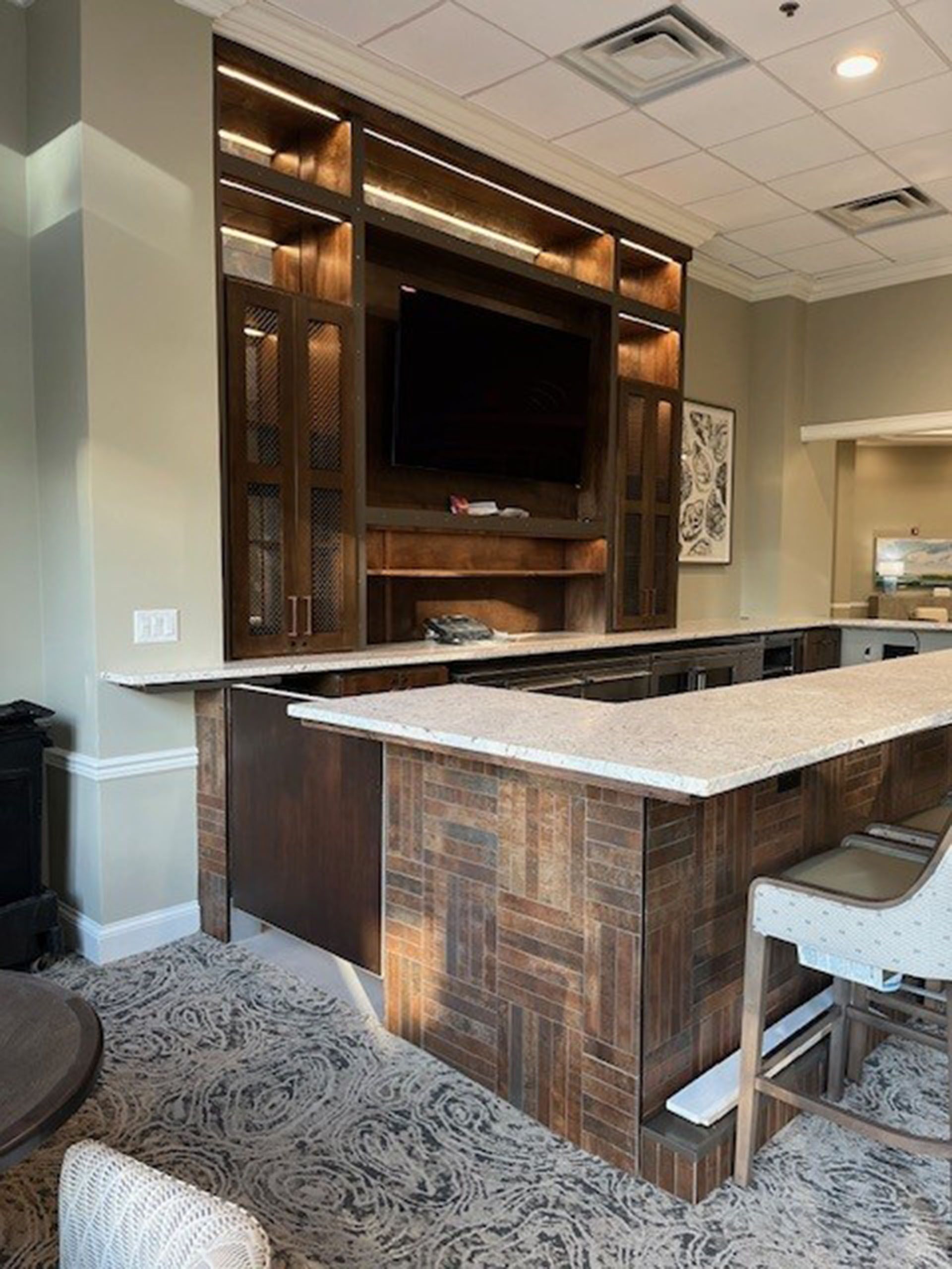 Commercial bar remodel - Built-in wooden bar with TV and shelving. Features light stone countertops, a patterned base, and recessed lighting.