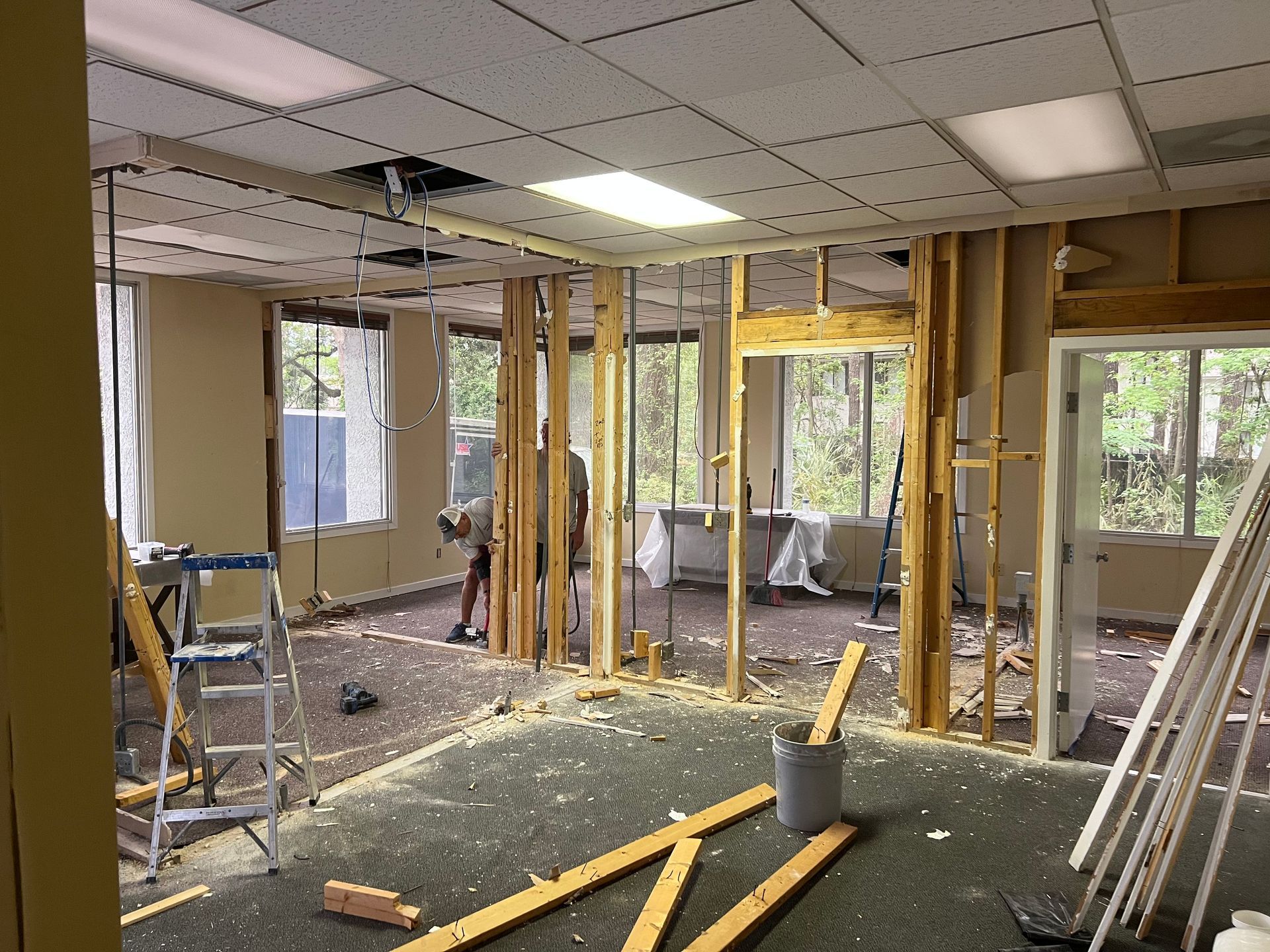 Interior room under construction, exposed studs, debris on the floor, windows with trees visible.