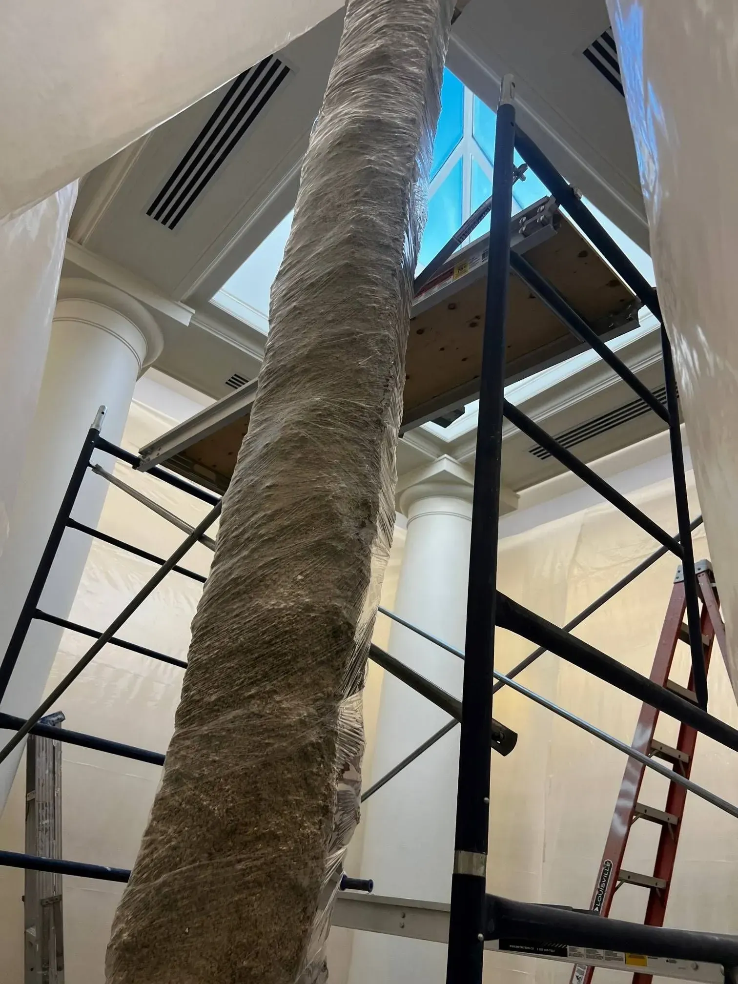 Column wrapped in protective material, scaffolding, and ladder inside a building.
