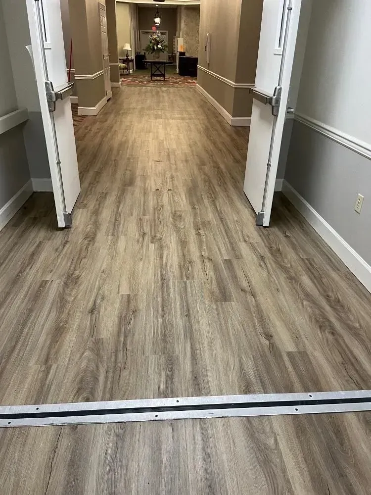 Hallway with light wood-look flooring, two open white doors, leading to a room with a table and flowers.