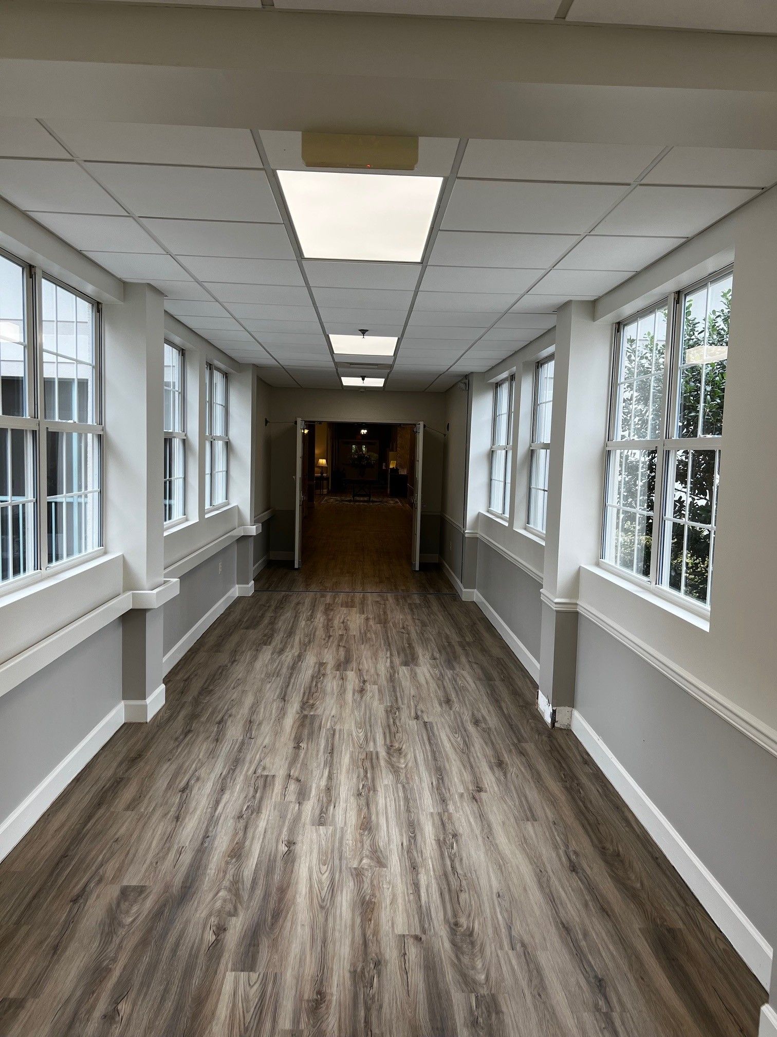 Long hallway with windows on both sides, wood-look floor, gray and white walls, leading to a darker room.