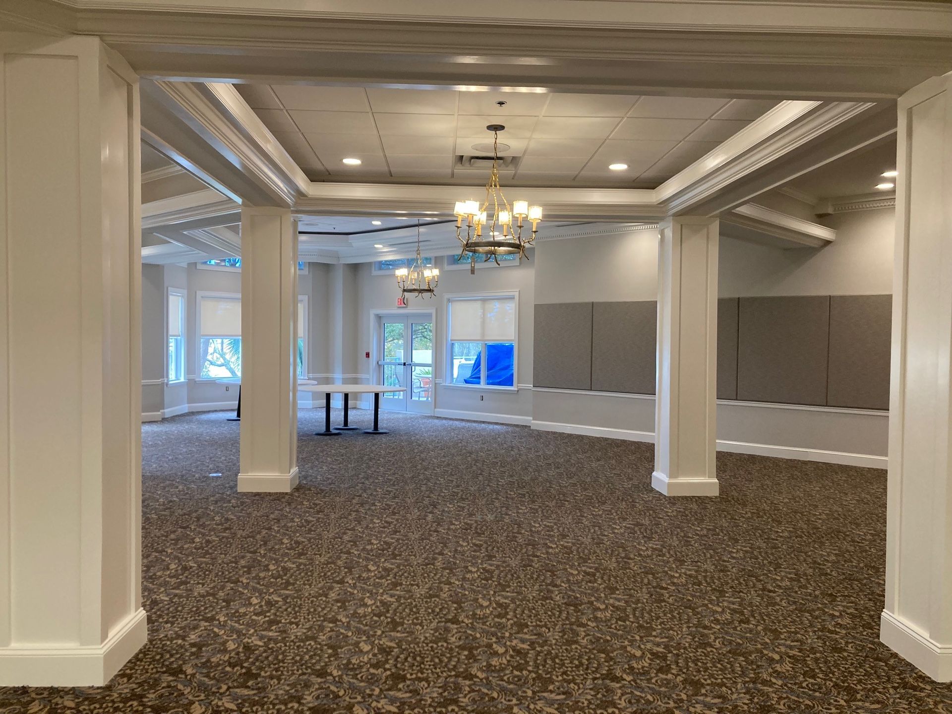 Empty event room with pillars, carpet, and a chandelier; neutral tones.