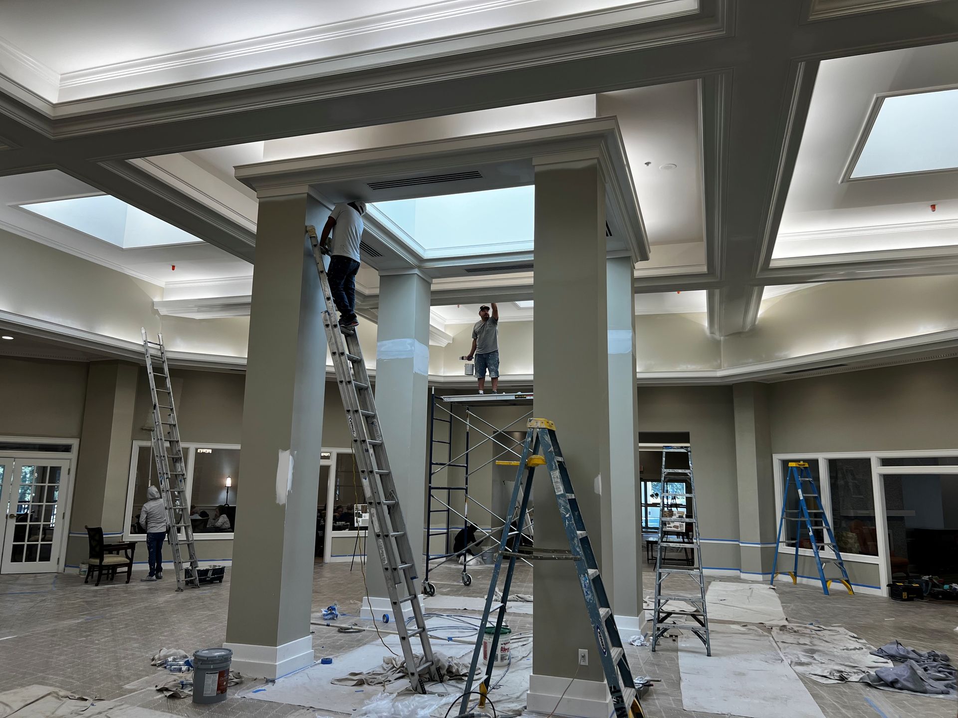People painting a building interior with columns, ladders, and paint supplies.