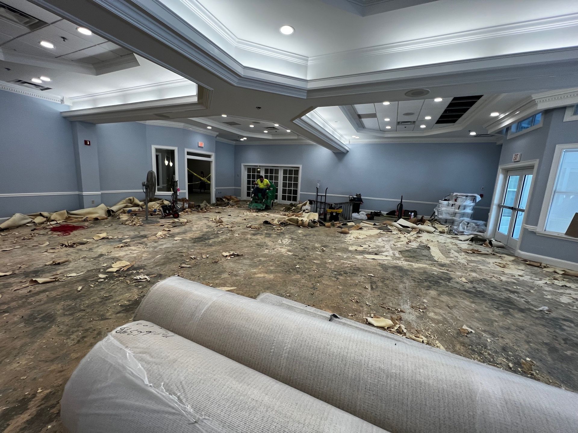 Renovation in progress: Room with exposed floor, debris, and rolled materials. Blue walls, white ceiling, and windows visible.