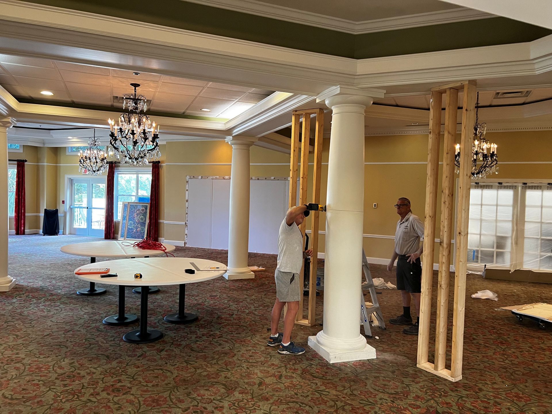 Two men building a wall inside a large room with chandeliers. Round tables are present.