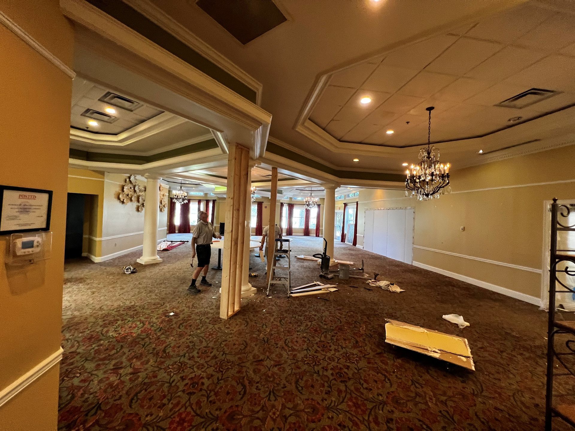 Interior of a room under construction with a person working. Beige walls, carpet flooring, pillars, and a chandelier.