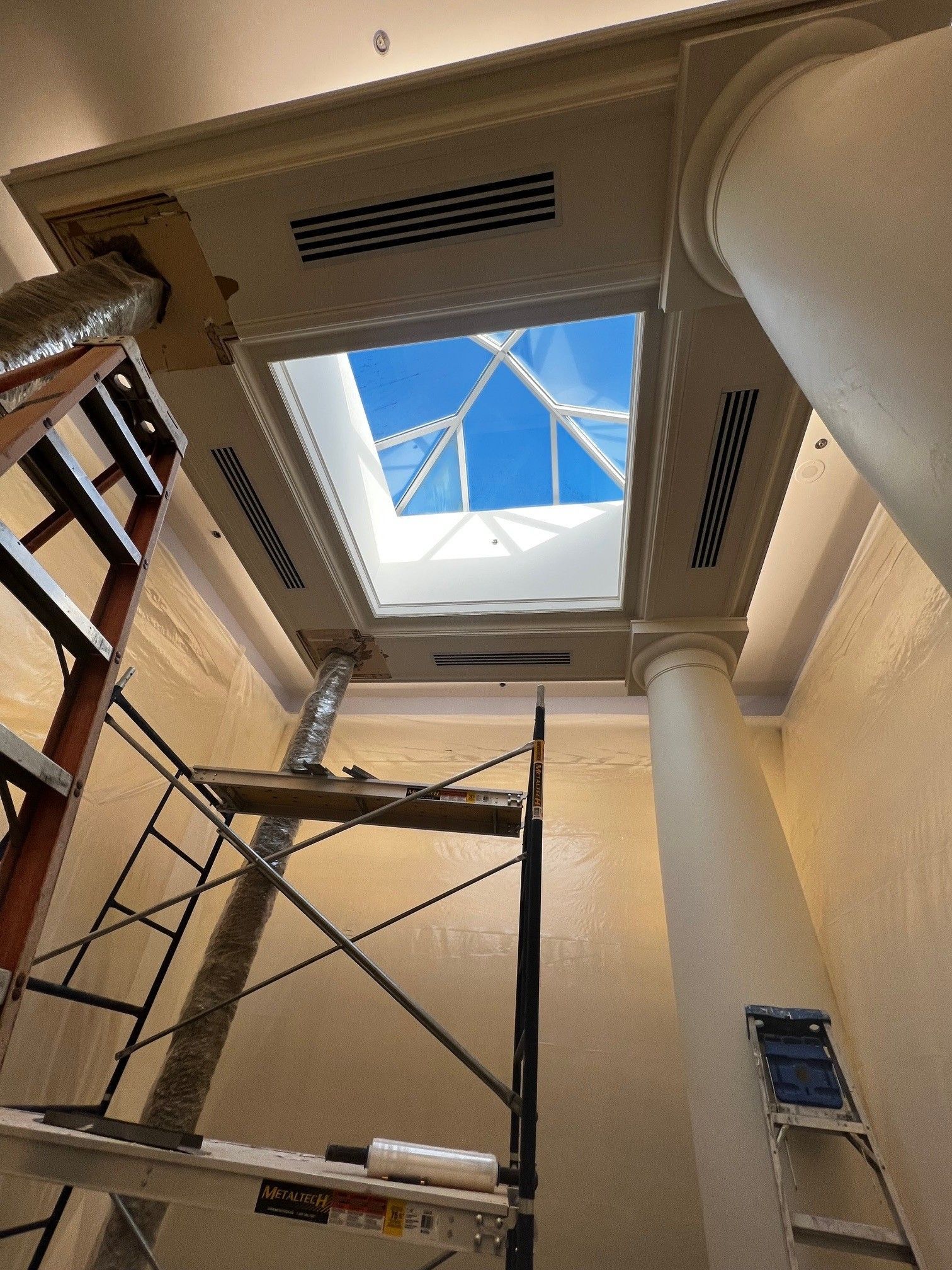 Interior view of a building with a skylight, a column, scaffolding, and a ladder. Blue sky visible through the skylight.