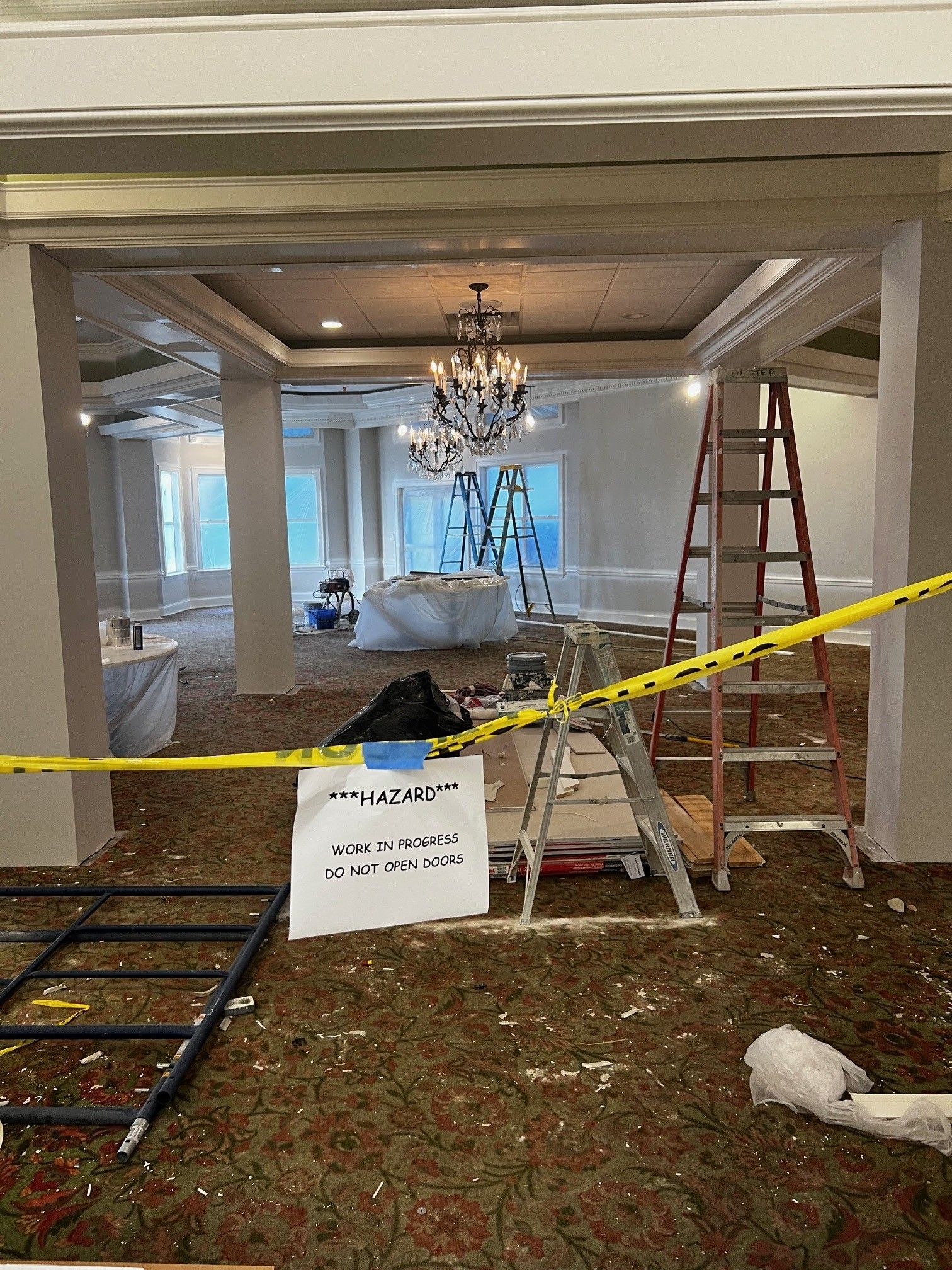 Room under renovation with chandeliers, ladders, and caution tape.