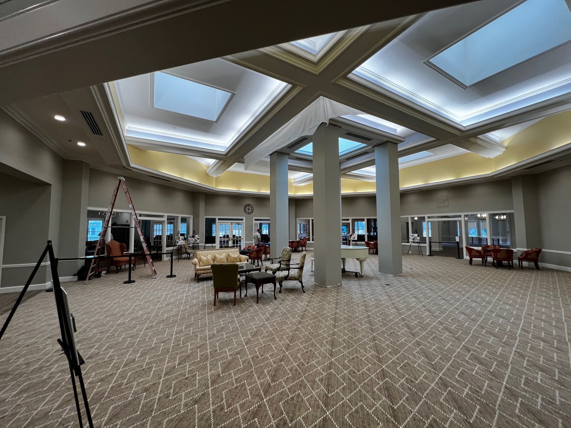Spacious lobby with carpet, seating areas, and skylights. Several people are present; a camera on a tripod is visible.