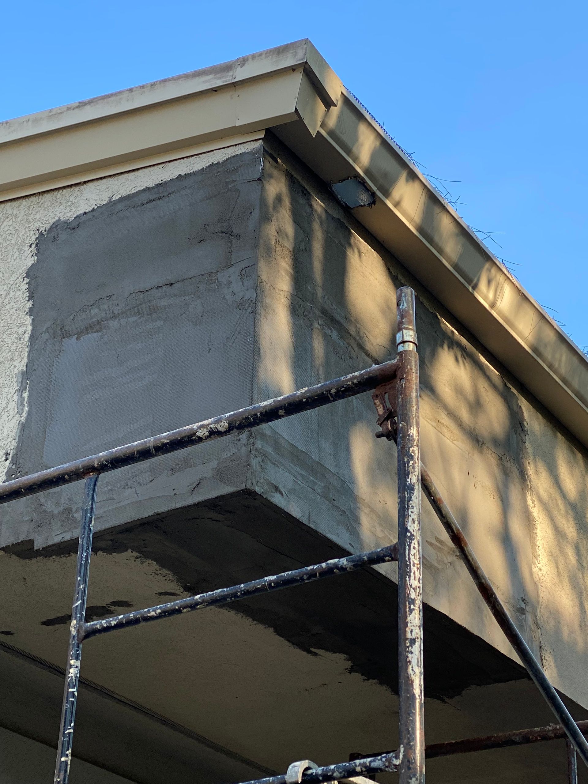 Exterior of a building with patched concrete, scaffolding visible beneath the eave, against a blue sky.