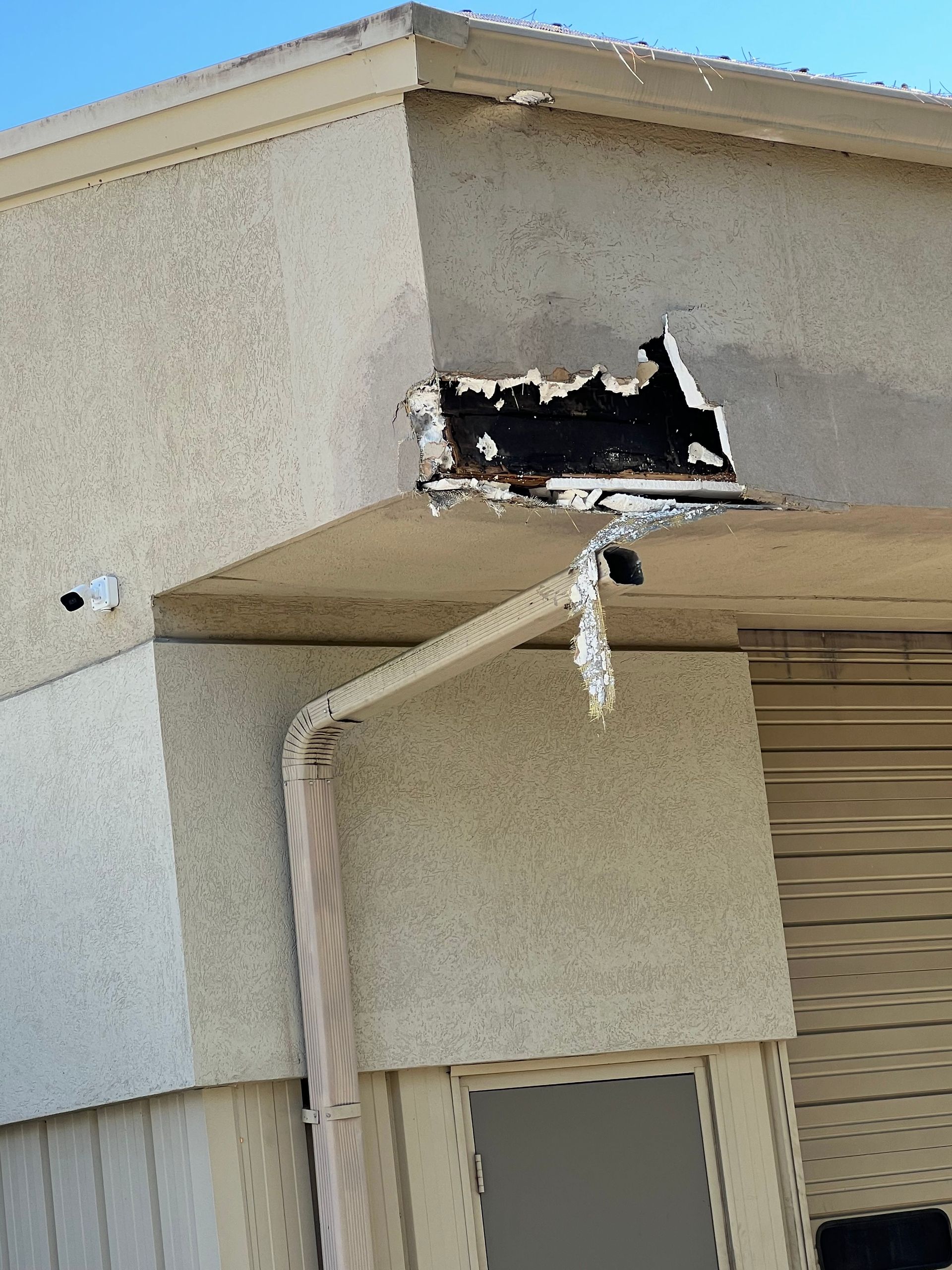 Damaged exterior building corner with peeling stucco, exposed black material, and a downspout.