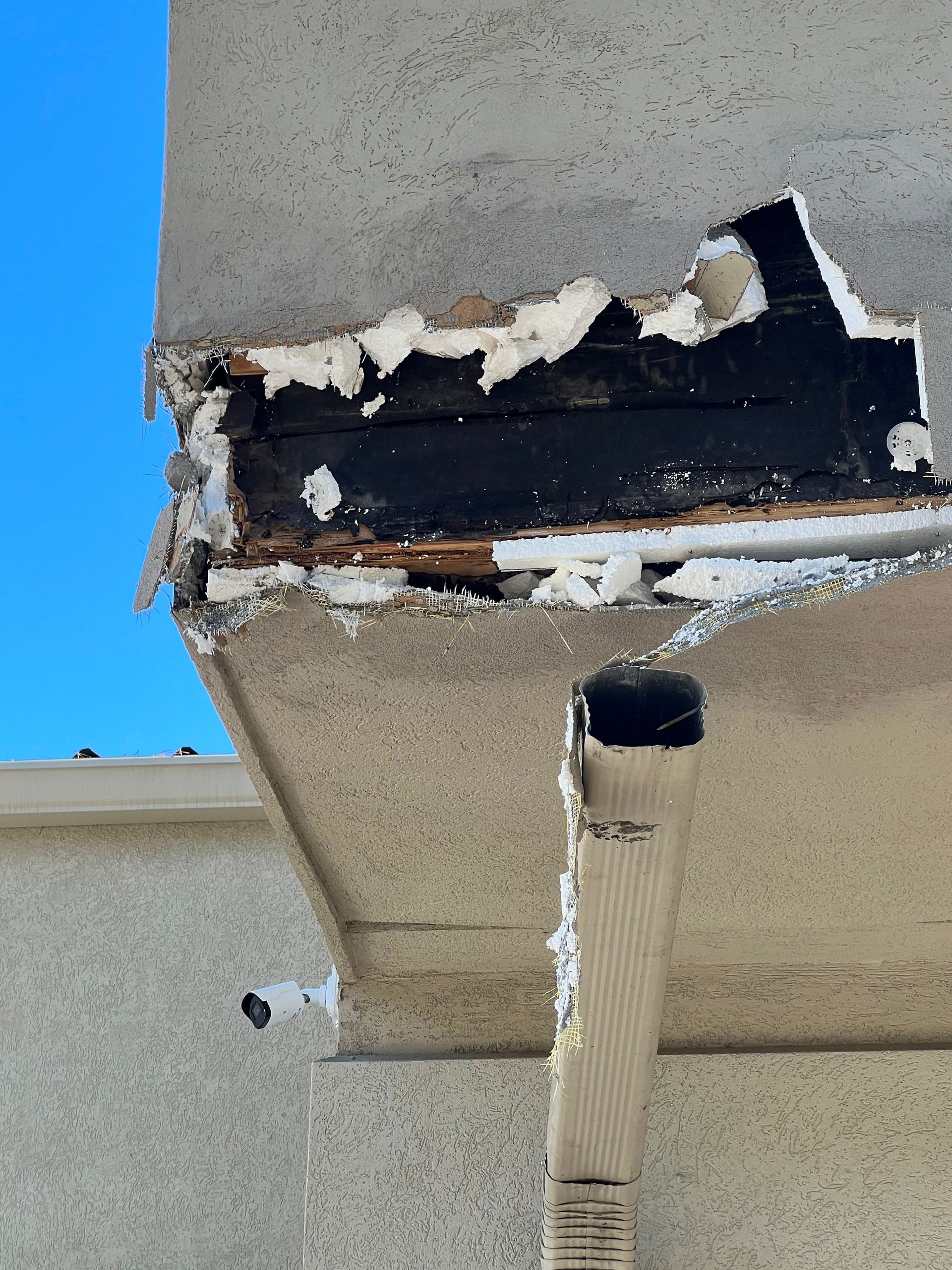Damaged exterior corner of a building, showing exposed wood and insulation against a blue sky.