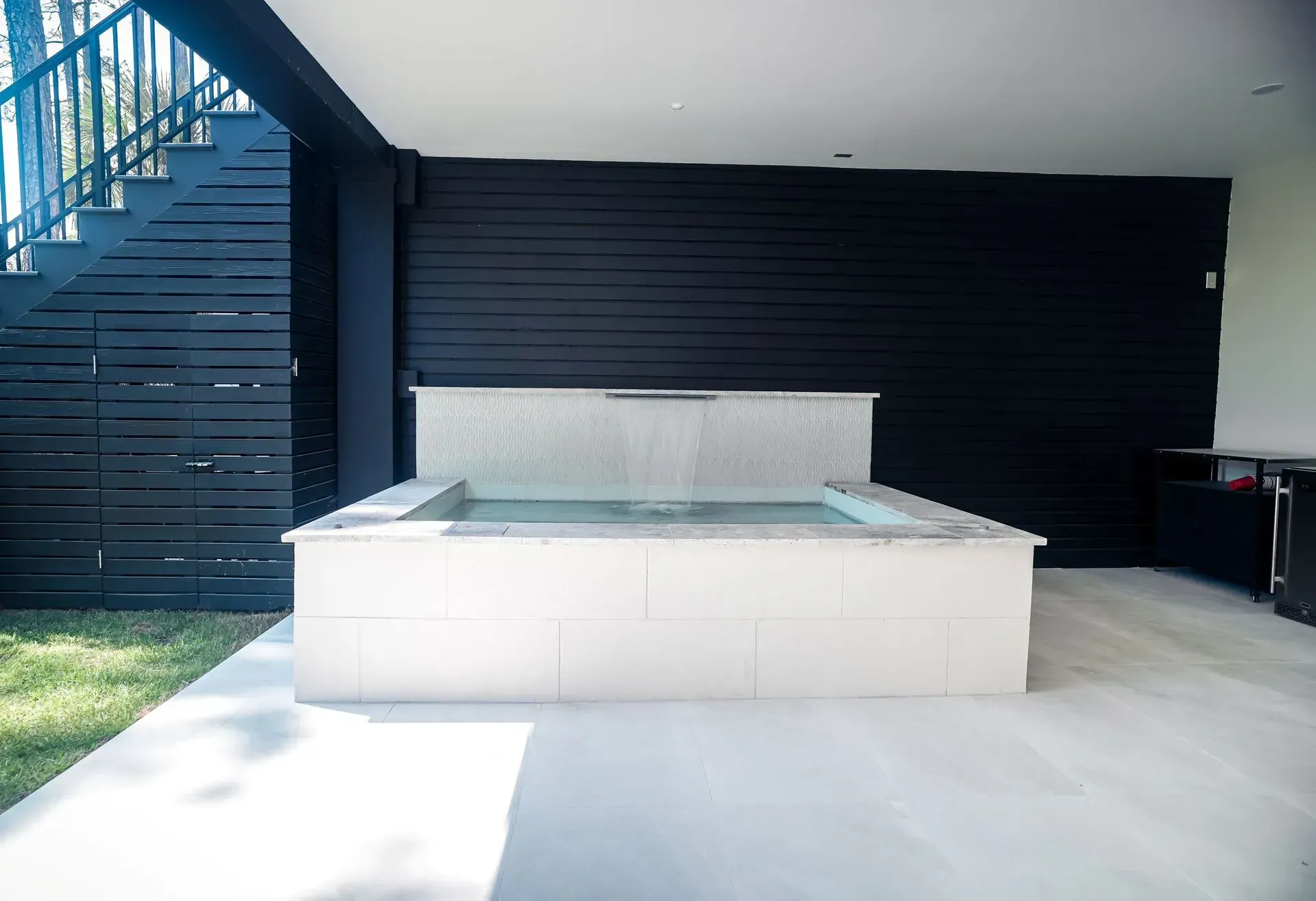 White hot tub under a black-painted wall. Steps and a grassy lawn are nearby.