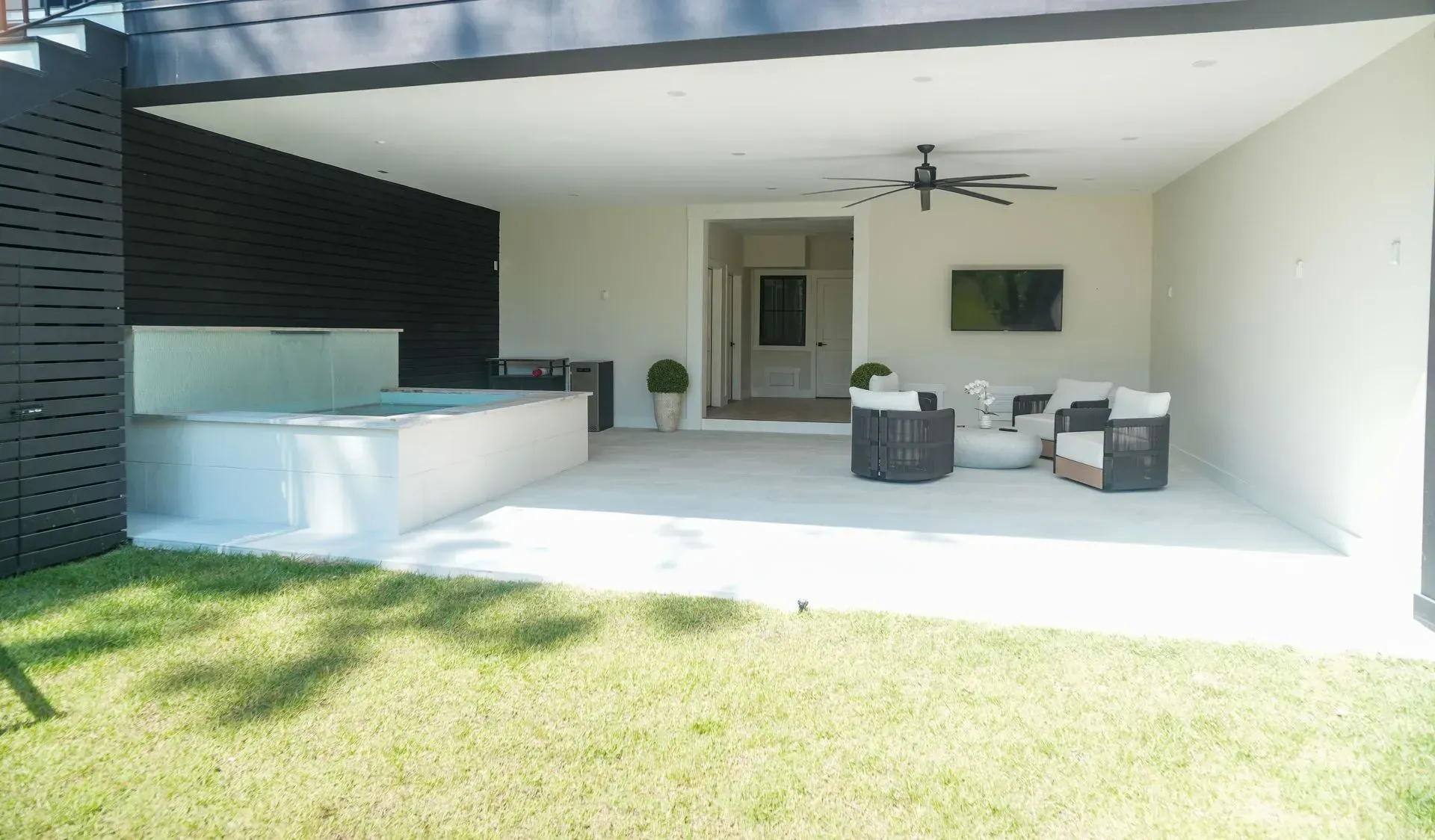 Outdoor patio with hot tub, seating, and mounted TV, on a concrete patio with a green lawn.