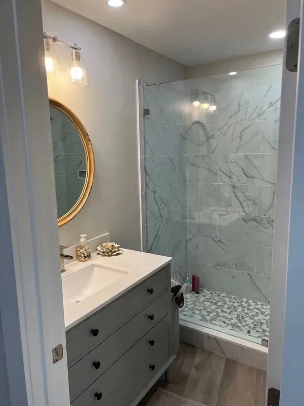 Bathroom with gray vanity, marble-tiled shower, round mirror, and wood-look floor.