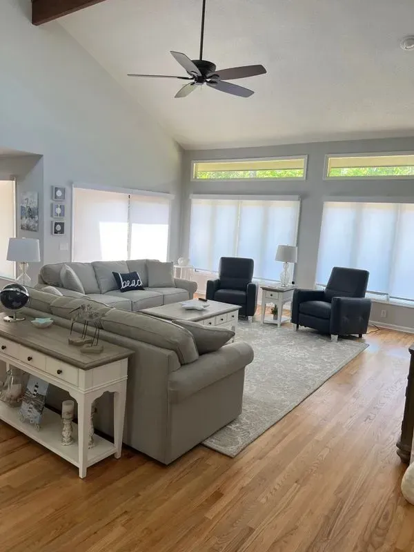 Living room with light gray sofa, two dark chairs, hardwood floors, and large windows.