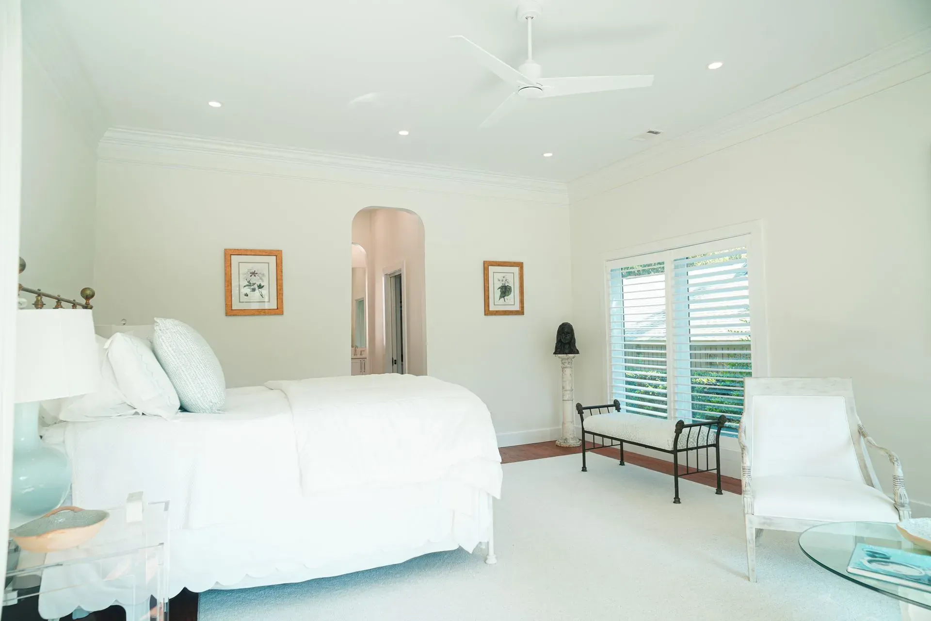 White bedroom with bed, artwork, seating, and shuttered window.