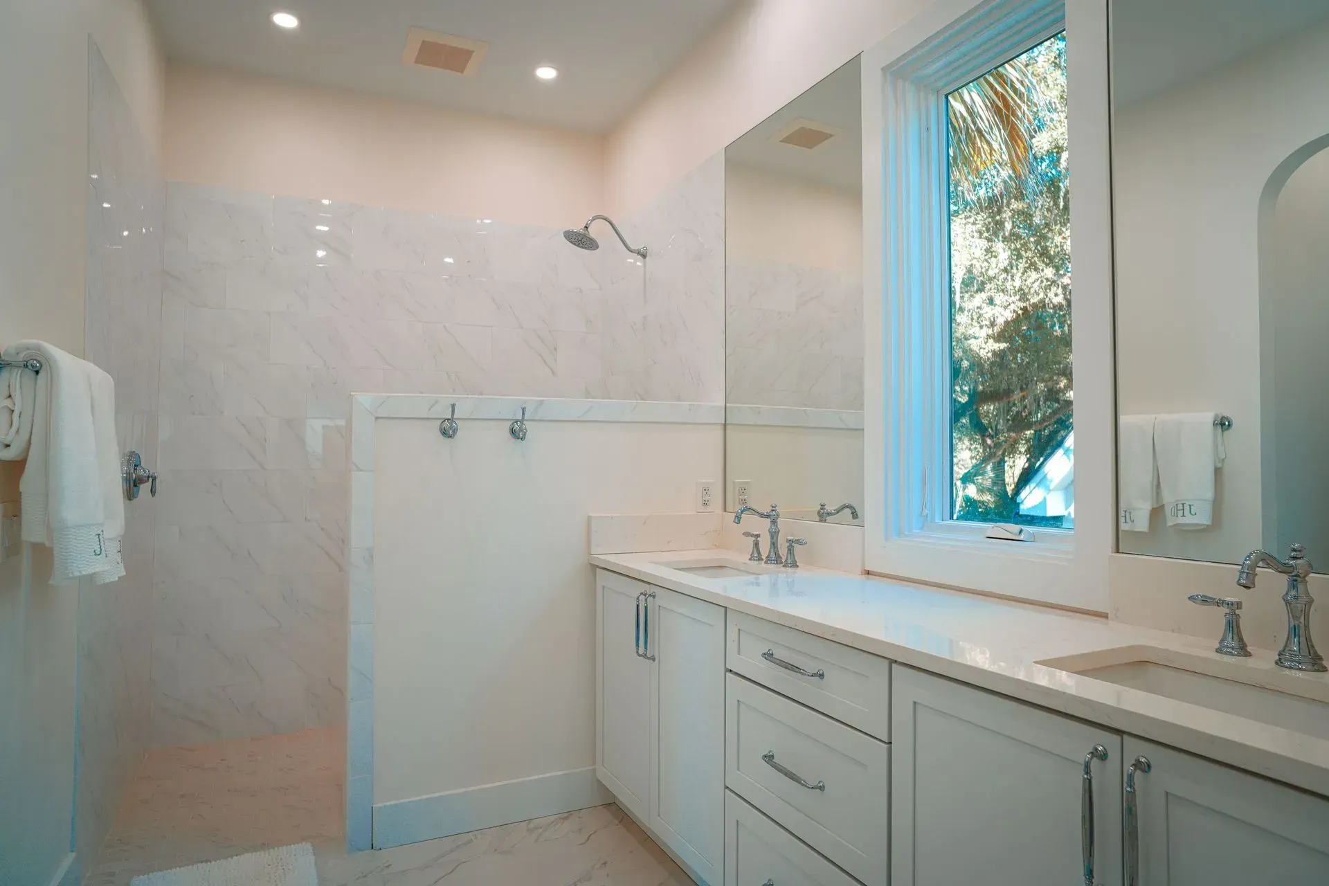 White bathroom with shower, double sinks, and a window with a tree view.
