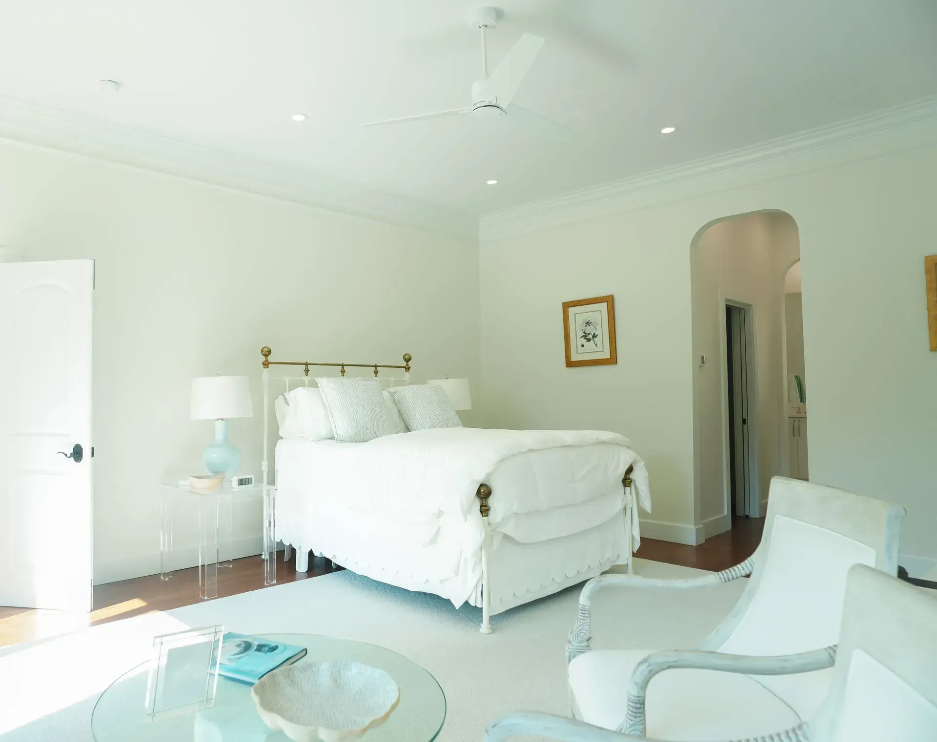 White bedroom with bed, two chairs, small side table, and round glass coffee table.