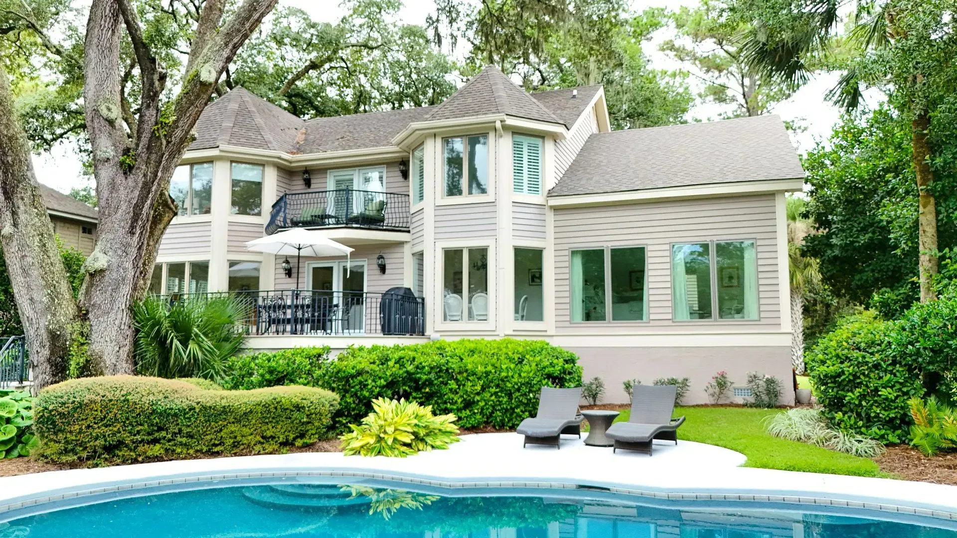Two-story house with a pool and outdoor seating. Light siding, green bushes, and a blue pool.
