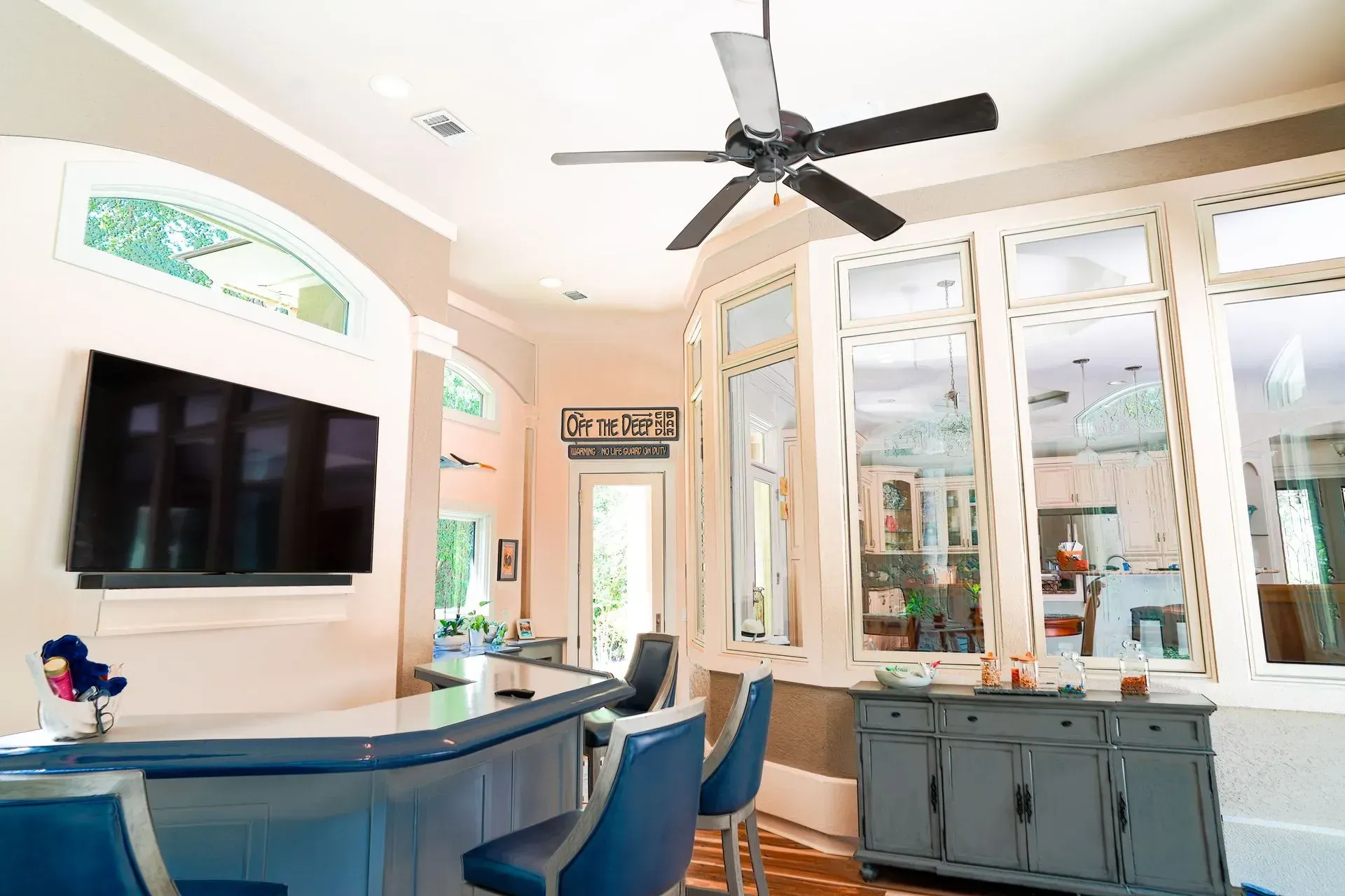Bar area with blue bar, stools, and wall-mounted TV. Large windows and a ceiling fan are present.