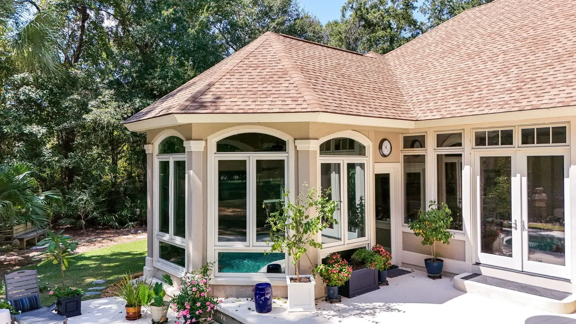 Exterior view of a house with large windows, tan walls, and a brown roof, surrounded by greenery.