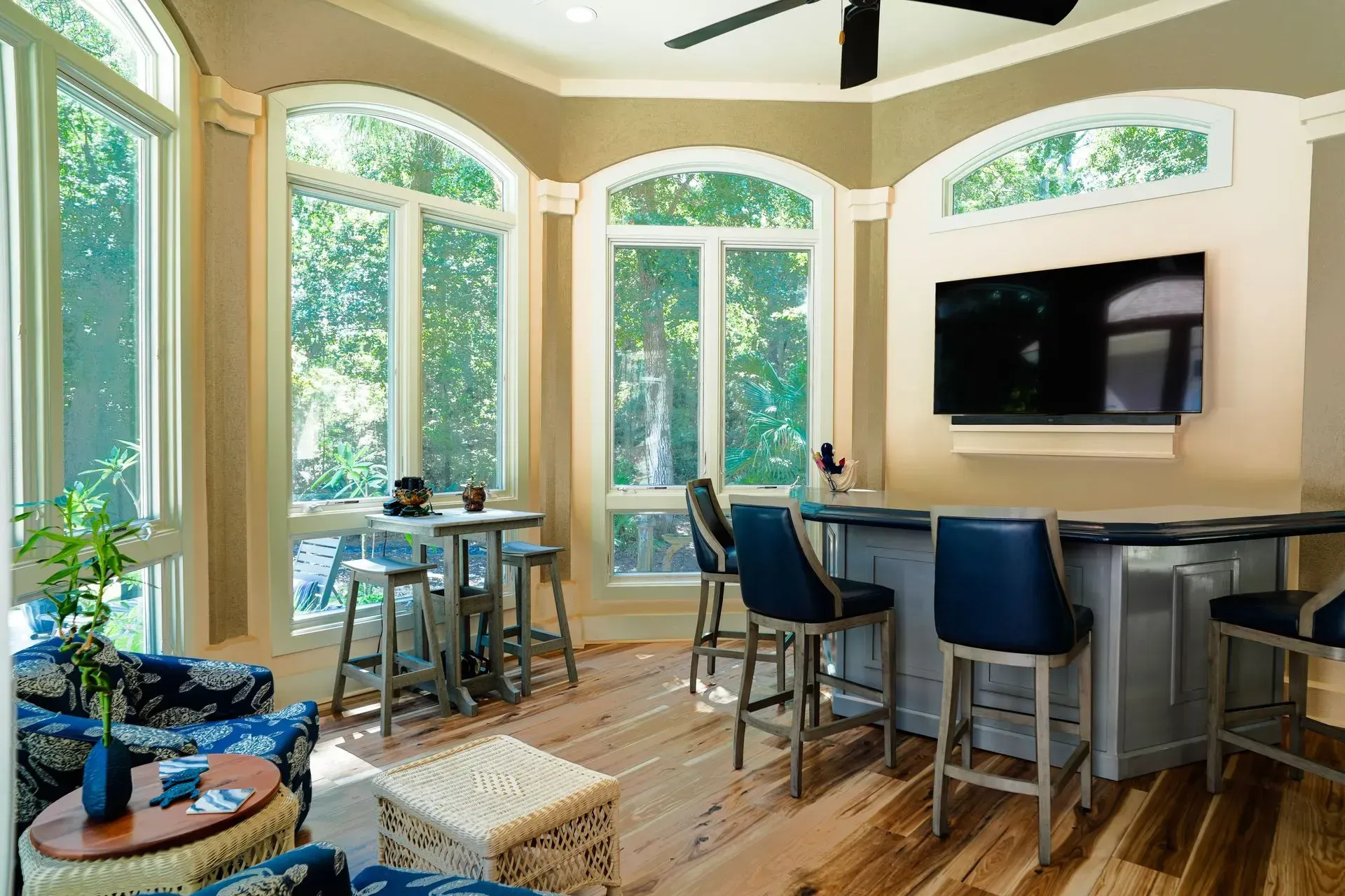 Bright room with windows, a bar with blue stools, and a small table with chairs; wooden floors and beige walls.