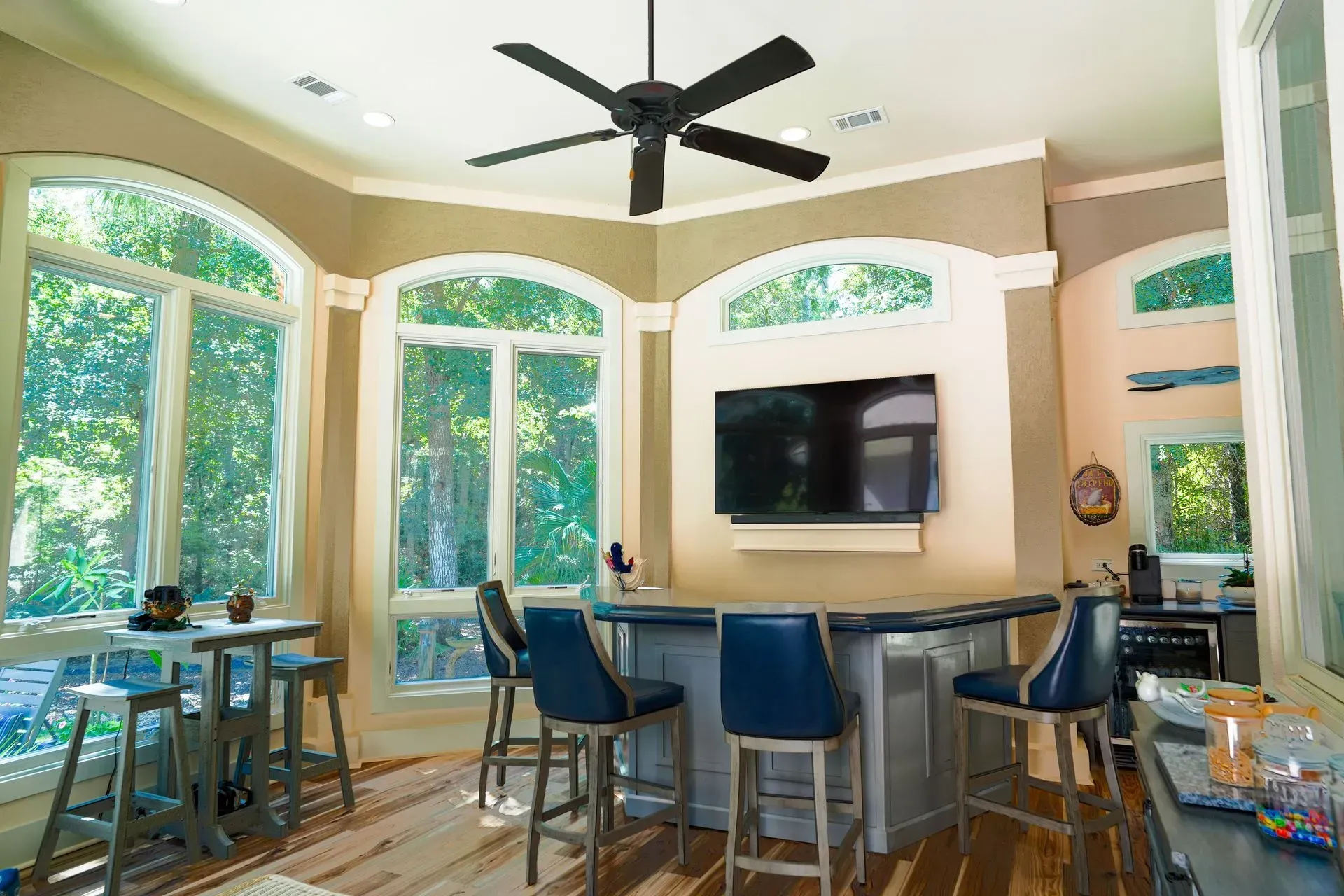 A bar area with blue chairs, a mounted TV, and large windows overlooking trees.