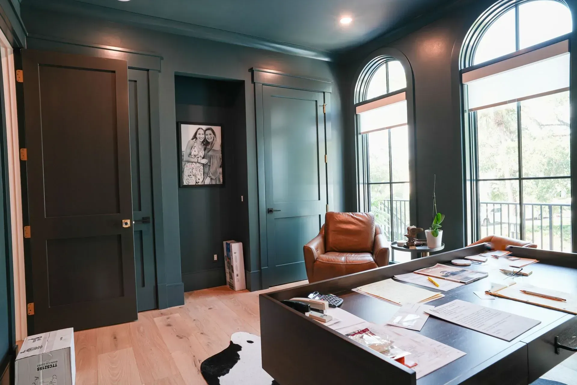 Room with dark green walls, arched windows, leather chair, wooden floor, and a large table.