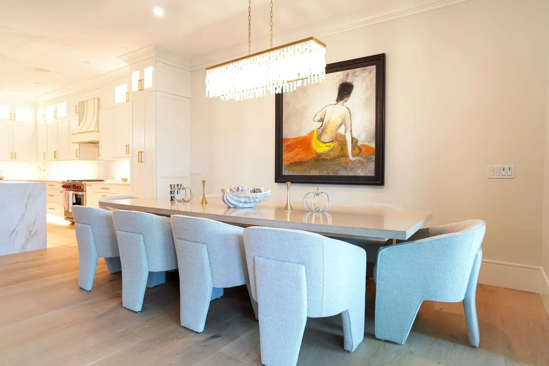 Dining room with long table, white chairs, and artwork. Overhead light fixture.