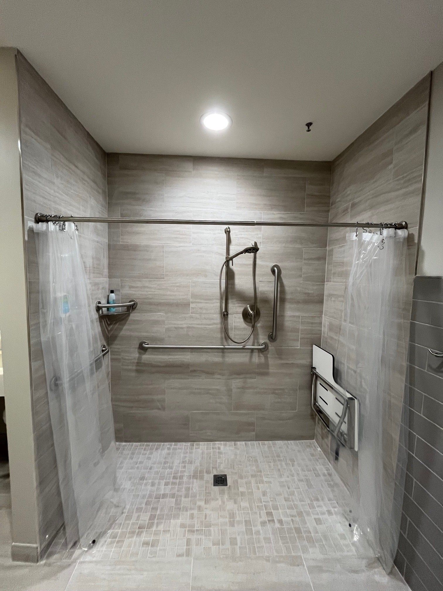 Accessible shower with grab bars and a fold-down seat. Light beige tile, two shower curtains.