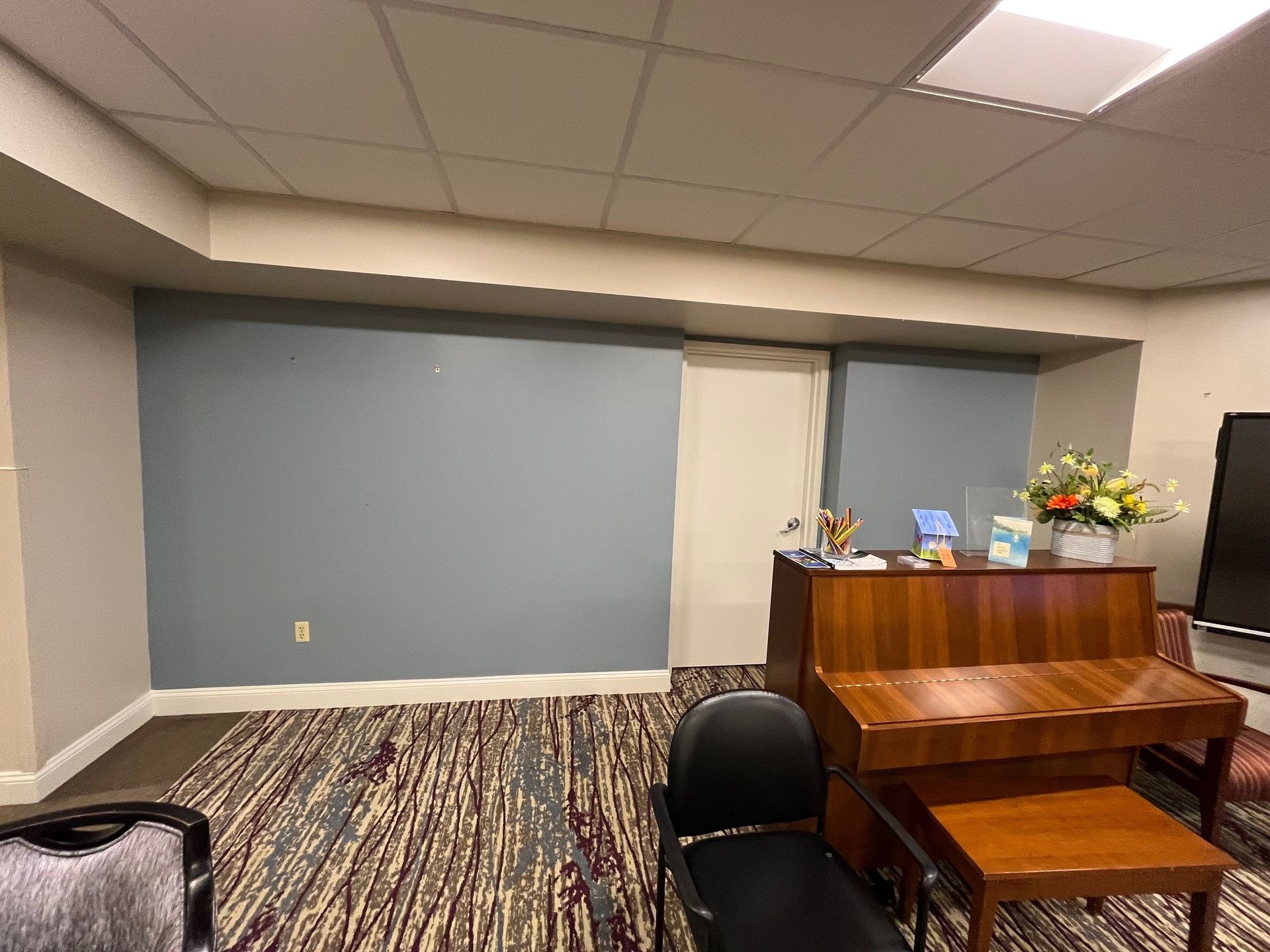 A room with a piano, blue wall, and patterned carpet. Desk with items, chair, and a door.