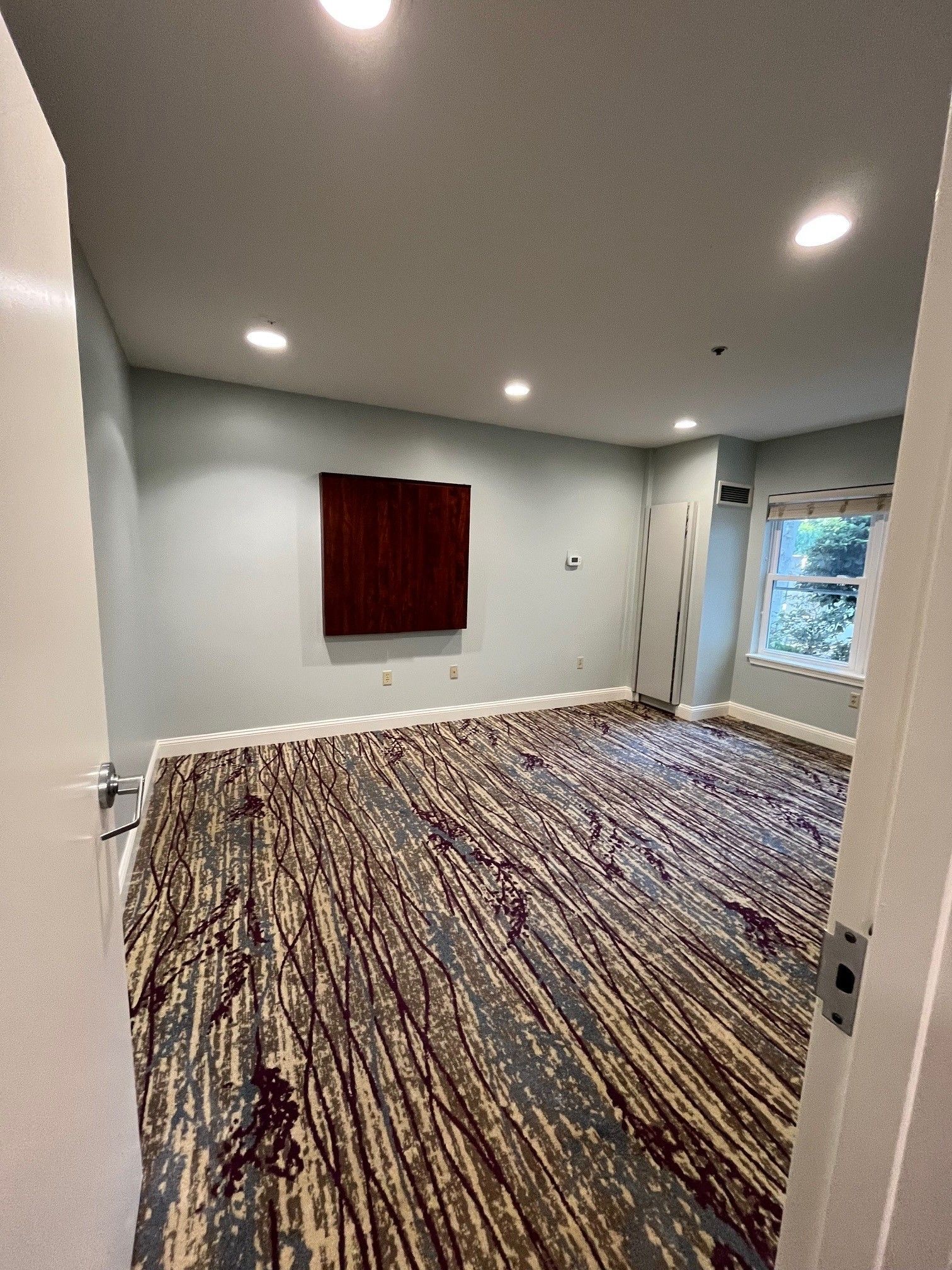 Empty room with patterned carpet, a window, and a dark artwork on a pale blue wall.
