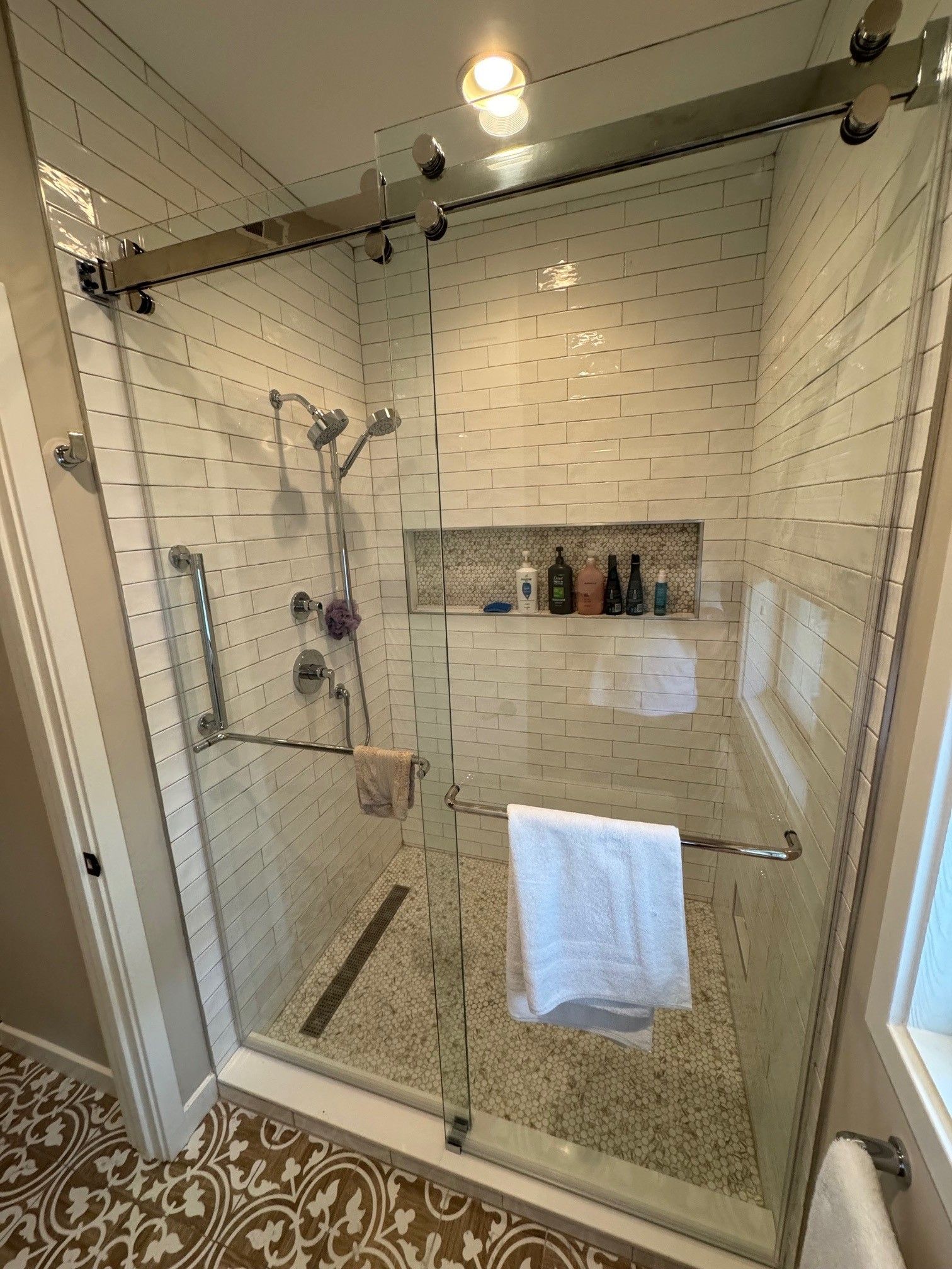 Shower with glass doors, white tiled walls, and pebble floor. Towels hang from the handles.