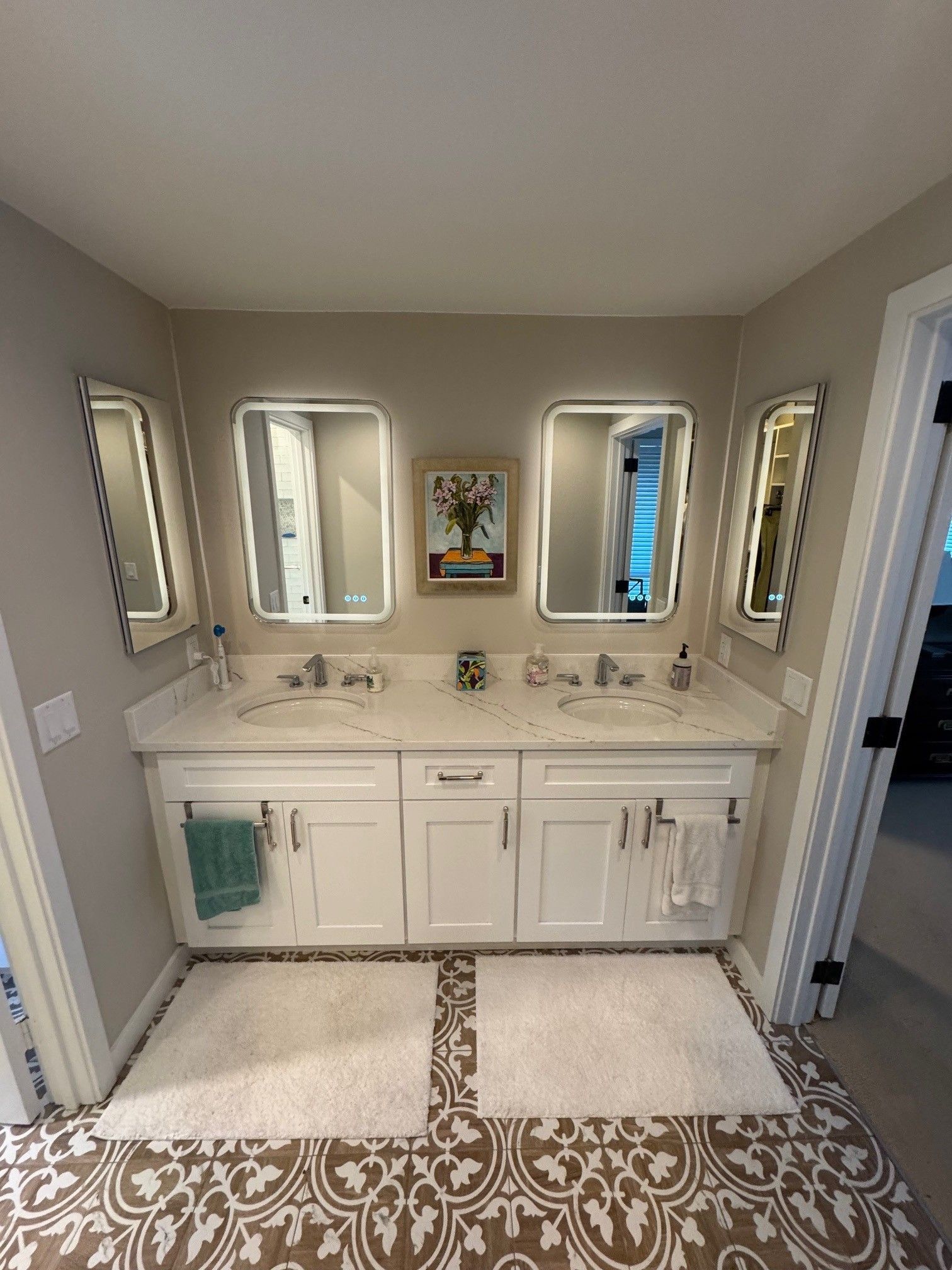 Bathroom with white double vanity, three mirrors, patterned floor, and two white rugs.