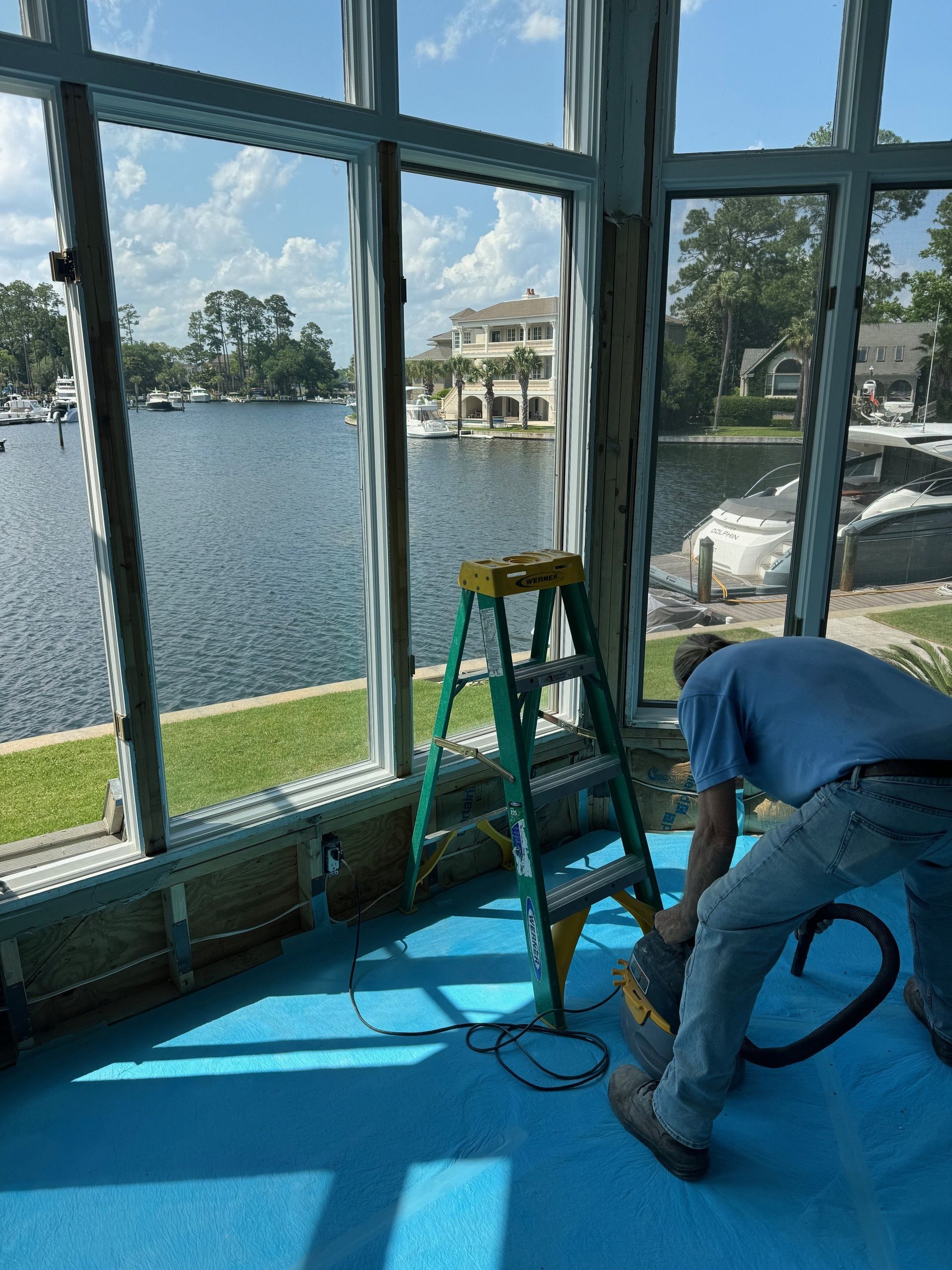 Person sanding inside a room with large windows overlooking water and boats, a ladder nearby.