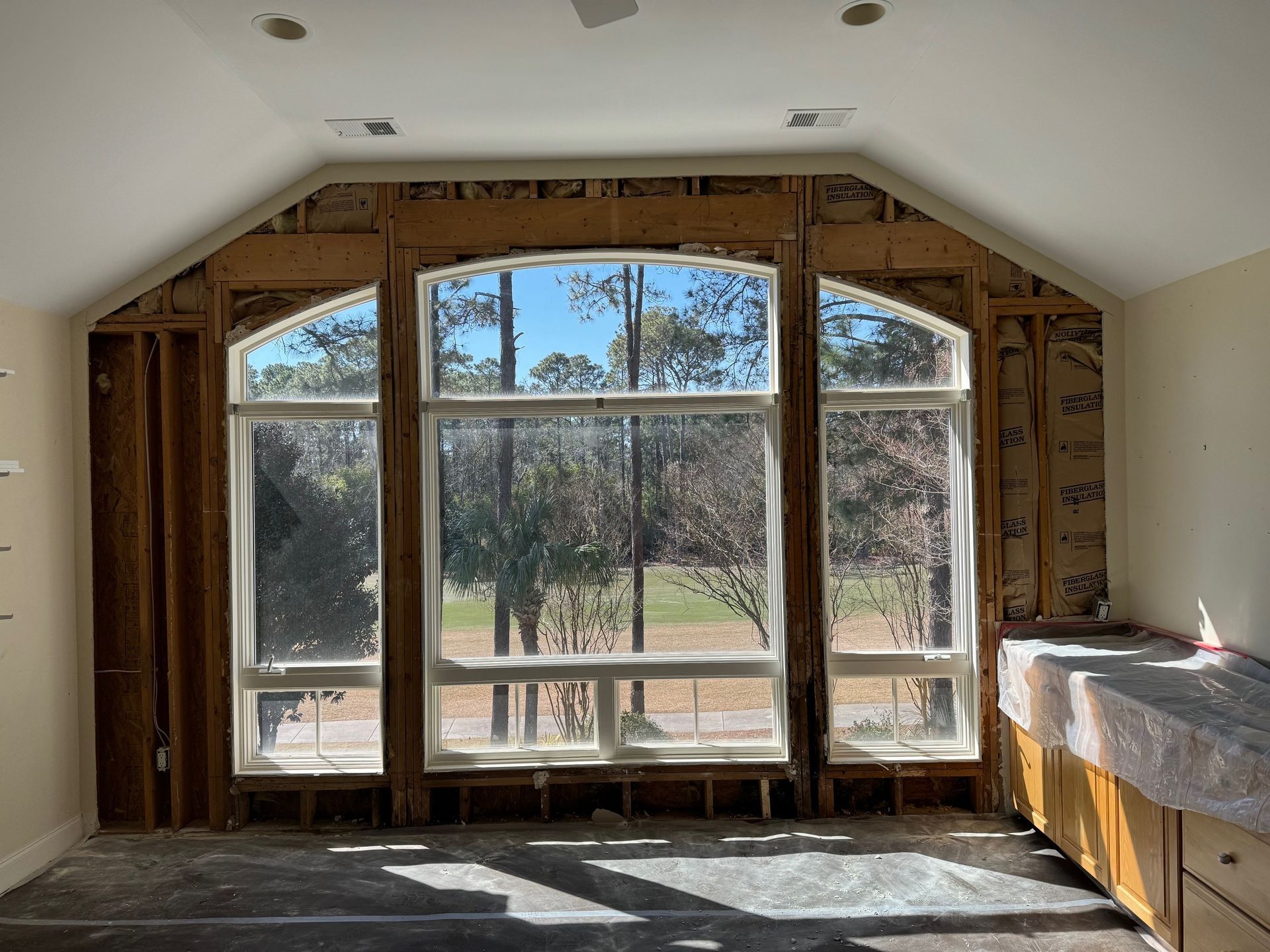 Interior view of a room under construction with a large arched window.  Exposed framing and insulation visible.