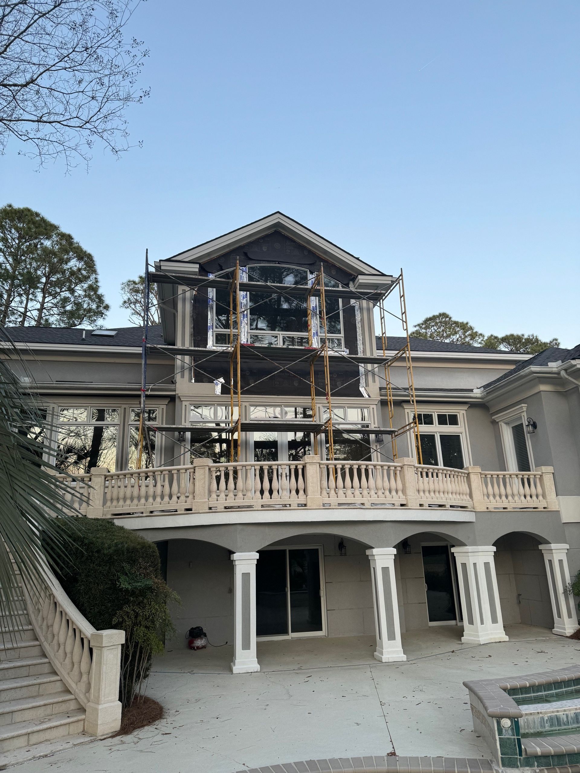 Exterior of a house under construction; scaffolding, windows, and balcony visible. Blue sky in background.