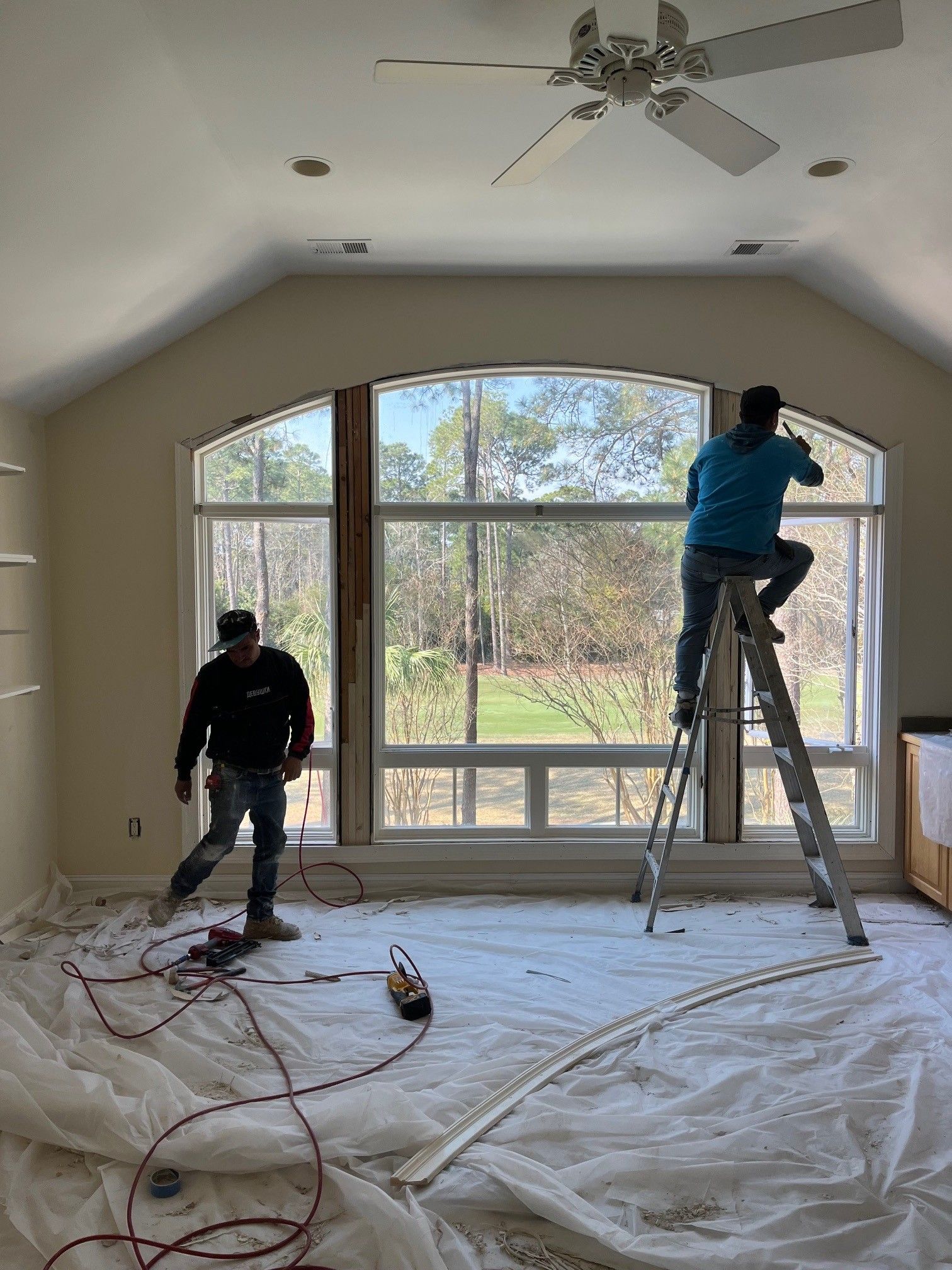 Two workers install a window, one on a ladder, in a room with a large arched window and covered floor.