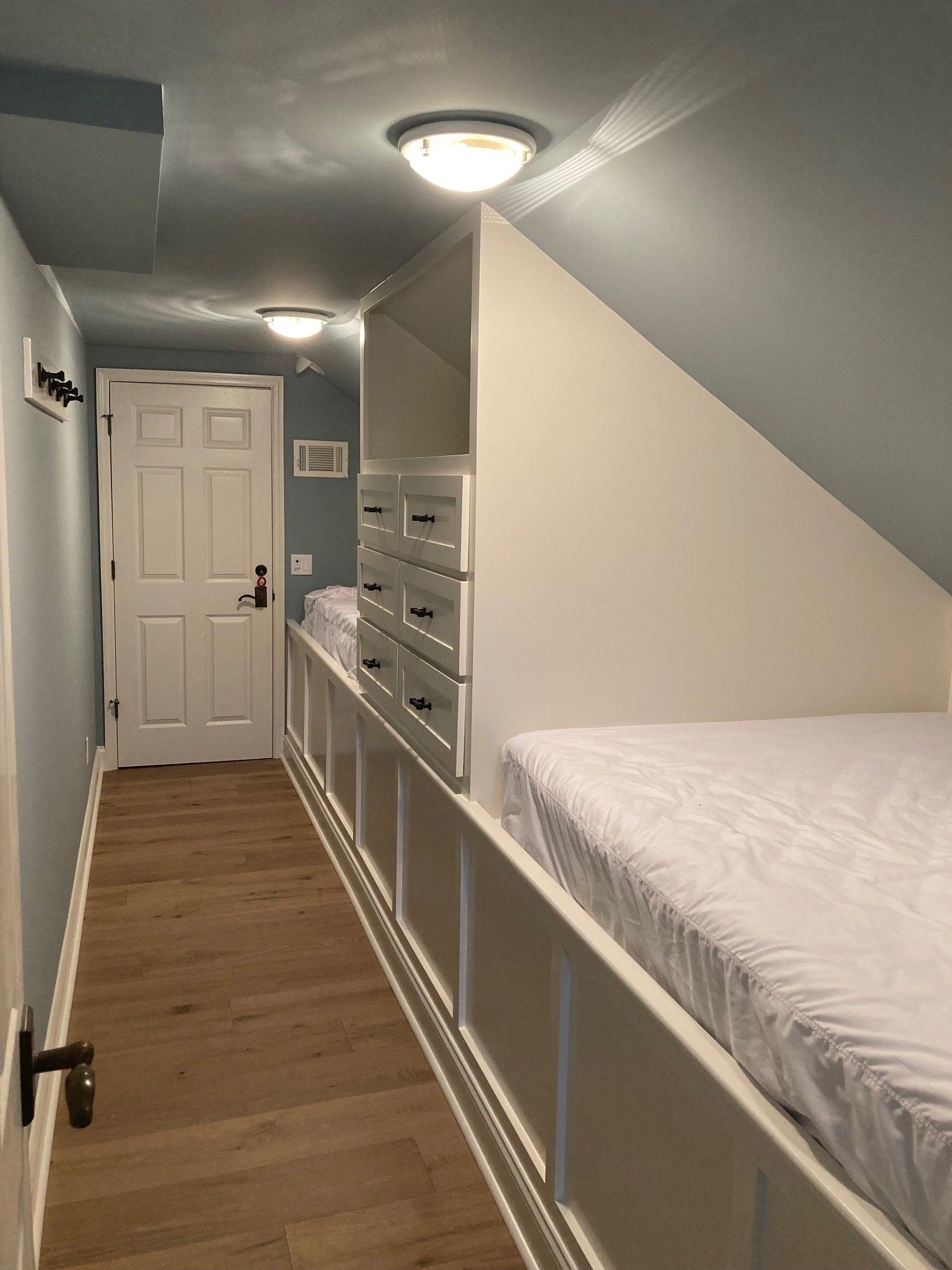 Narrow bedroom with built-in bed, drawers, and door. Blue walls, light wood floor, white bed. Two ceiling lights.
