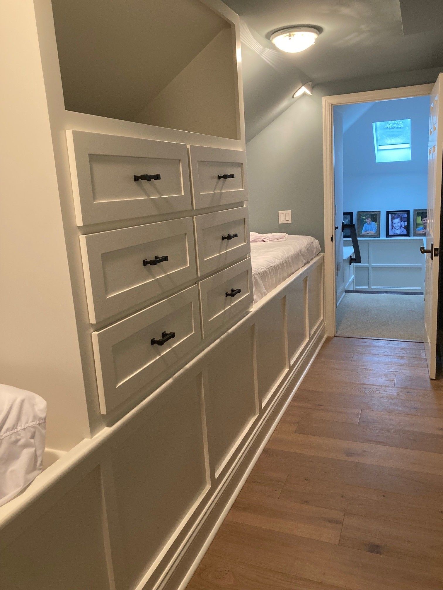 Built-in white storage and bed in hallway, drawers with black pulls, light blue wall, wood floor, door to other room.