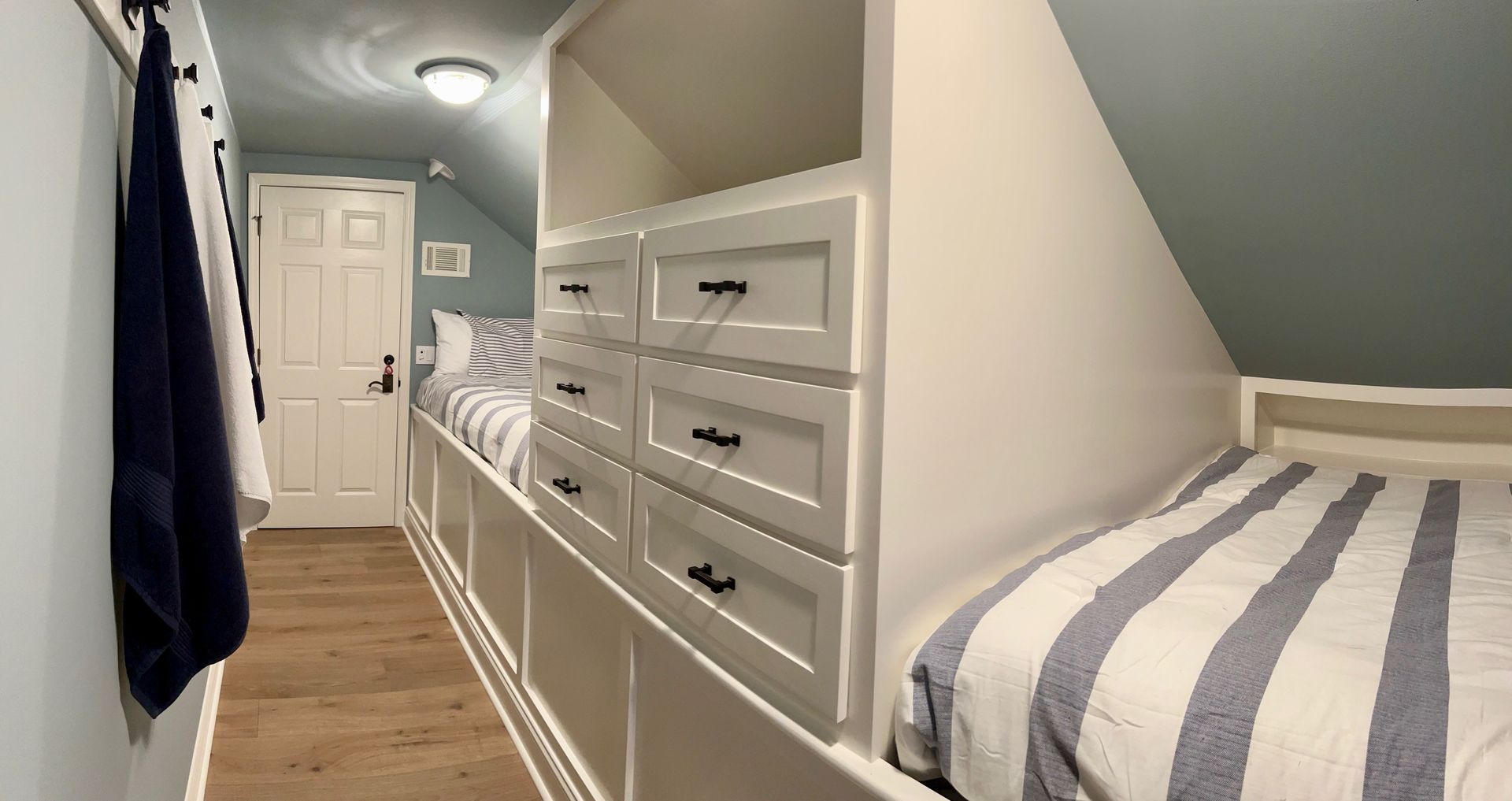 Built-in white cabinets and drawers with two beds against a sloped ceiling. Light wood floors, blue accents.
