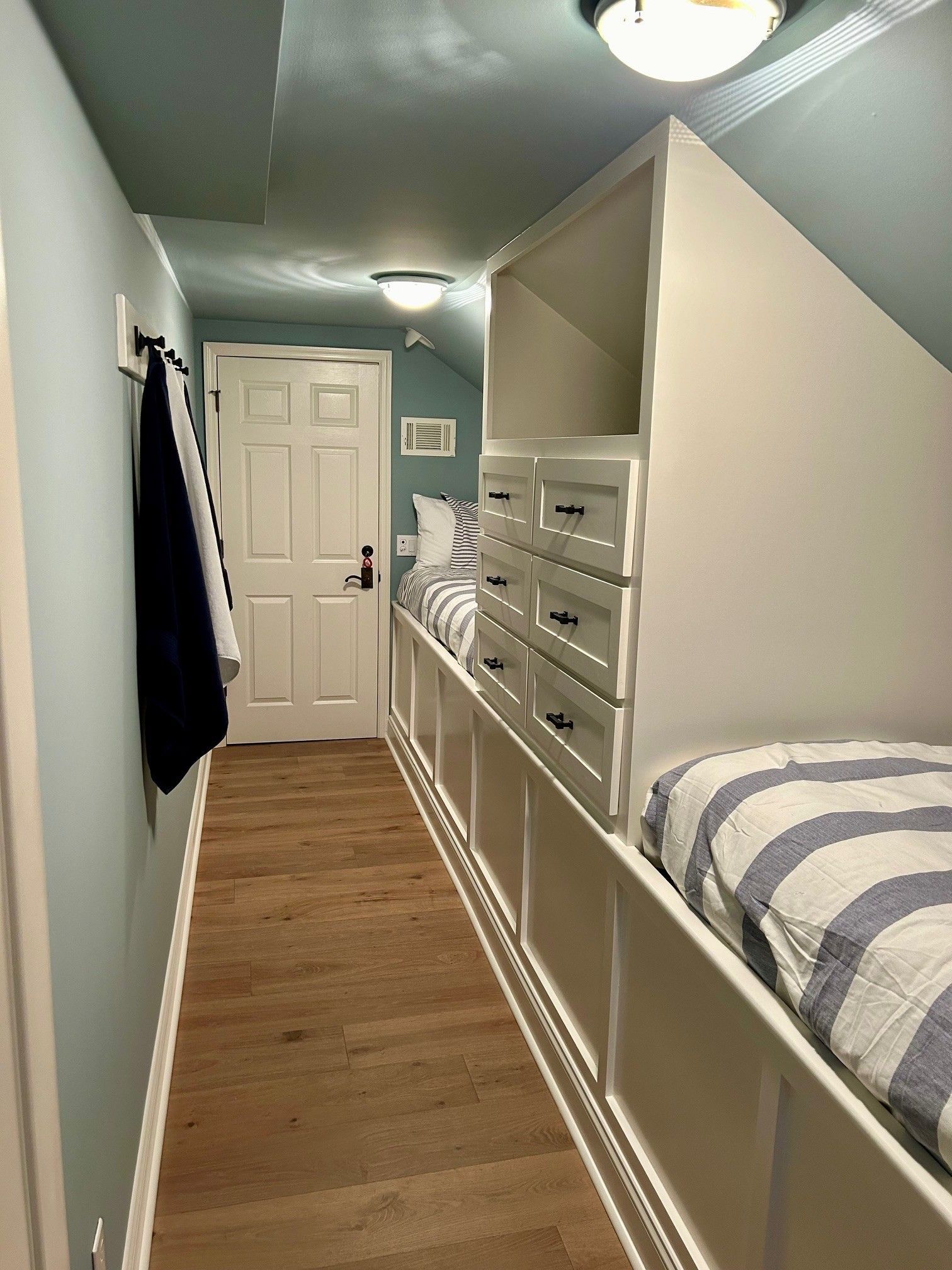Narrow bedroom with built-in bed, drawers, and door. Blue and white bedding, pale walls, light wood floor.