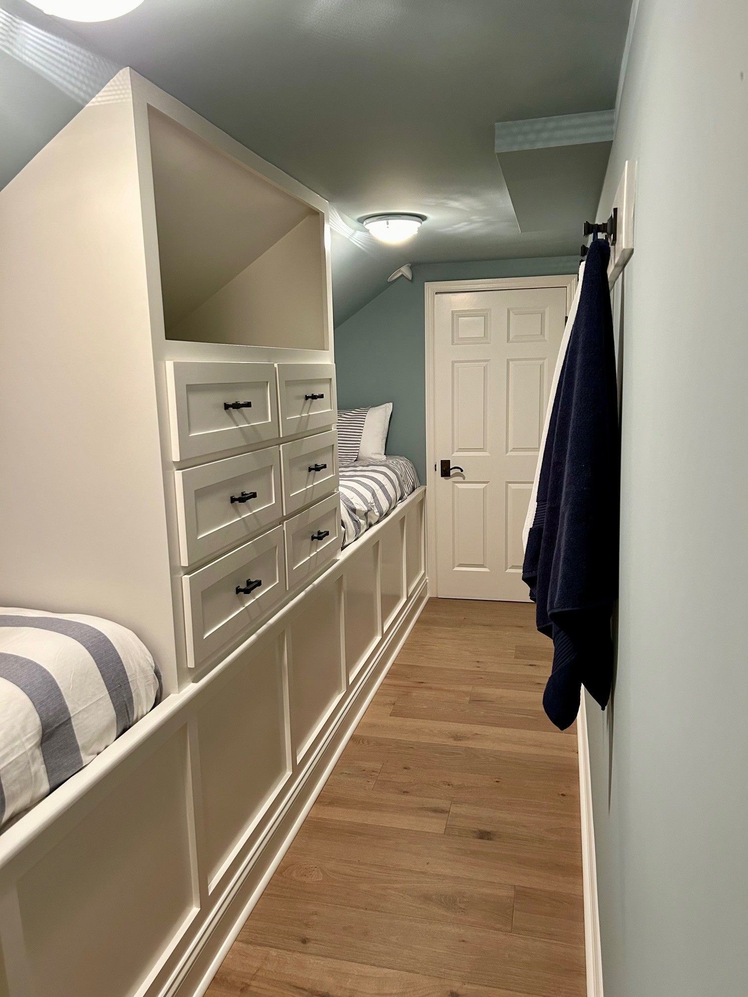 Built-in bunk beds with drawers in a narrow hallway with a door. Blue and white striped pillows on beds.